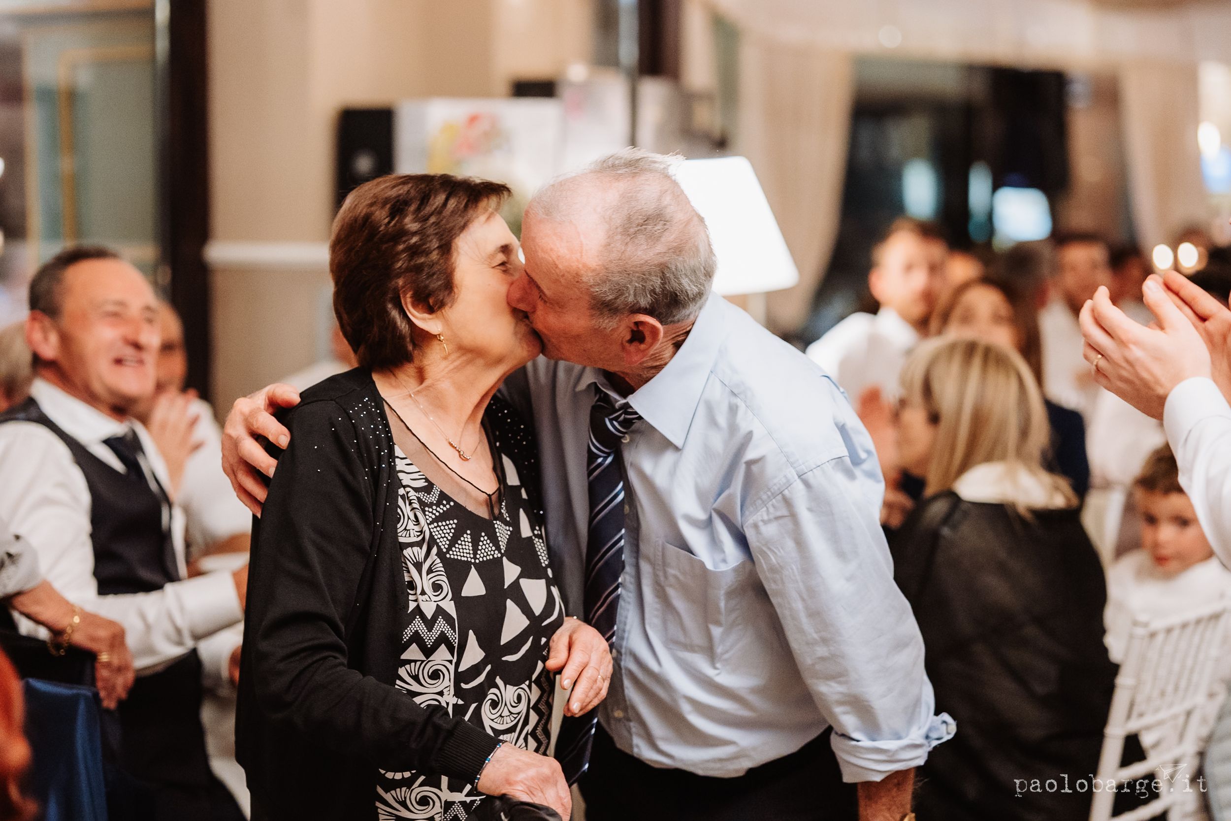 A couple shares a joyful moment dancing together at a celebration while others applaud in the background.