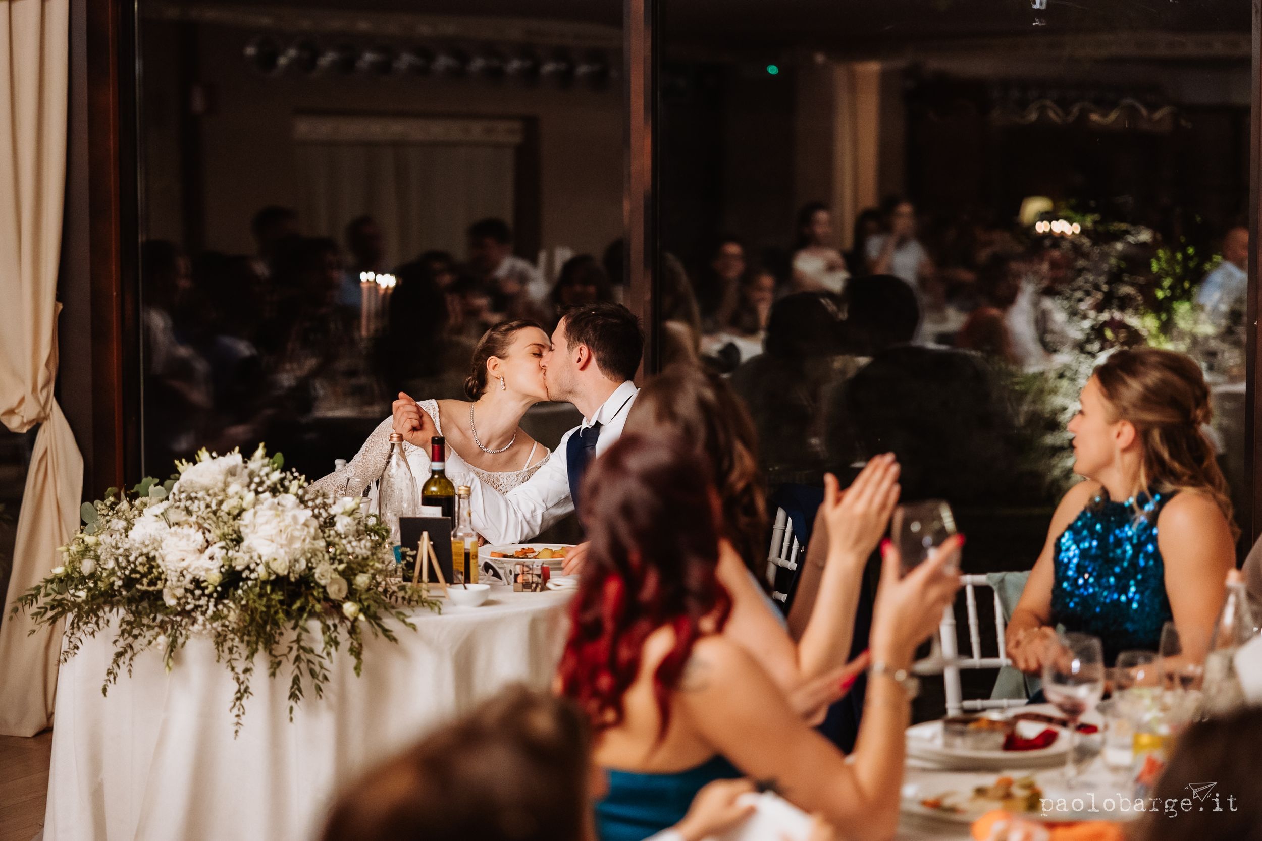 A couple sits at an elegant wedding reception head table with floral arrangements while guests applaud during dinner.