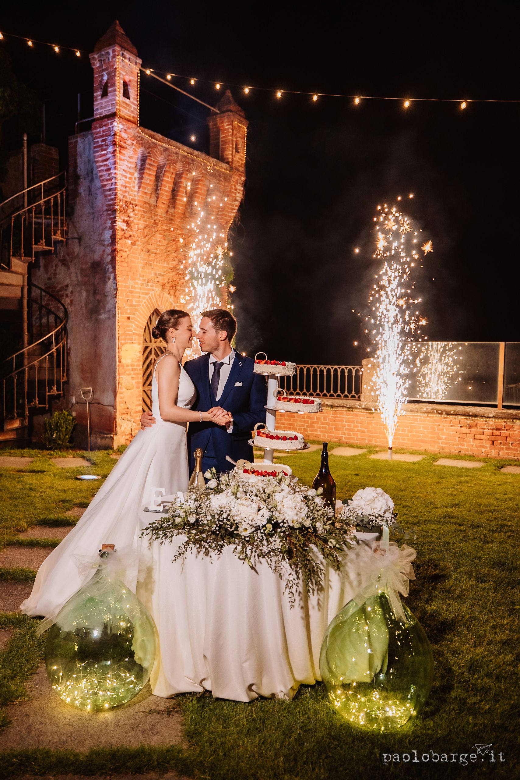 A newlywed couple celebrates at their evening reception with sparklers and string lights at a historic stone castle venue.