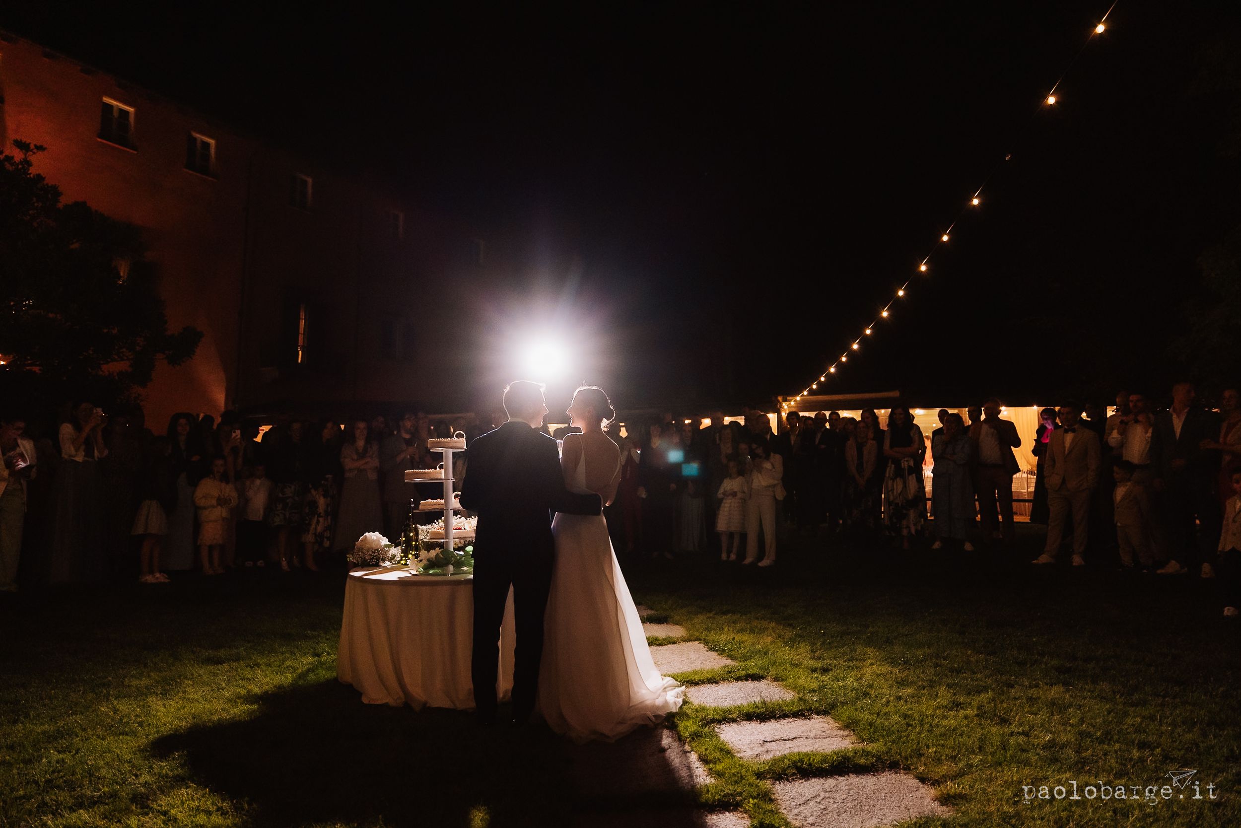 Couple embraces at nighttime outdoor wedding reception with string lights and guests in background.