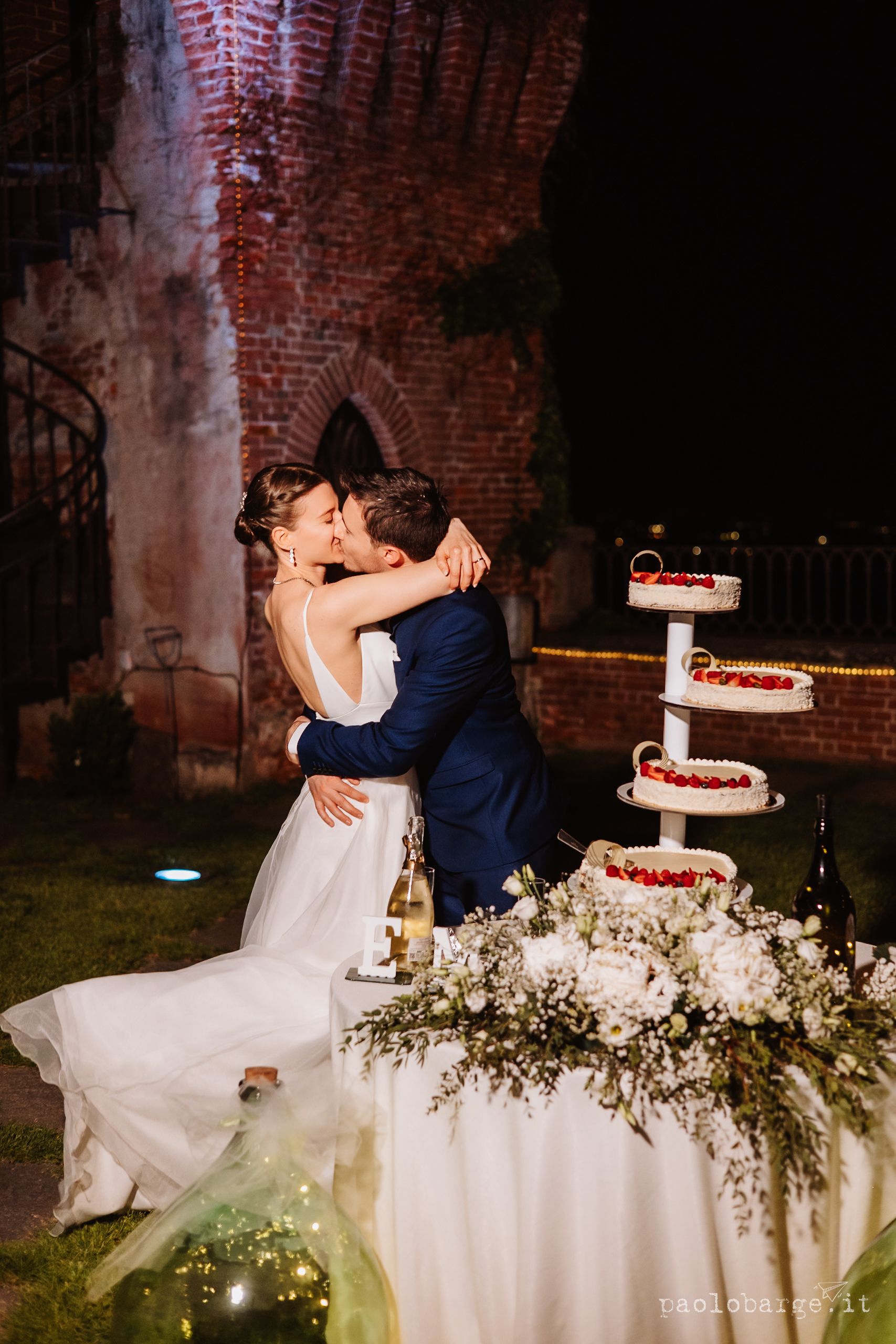 Couple shares romantic kiss during wedding reception by elegant multi-tiered cake adorned with red berries and florals.