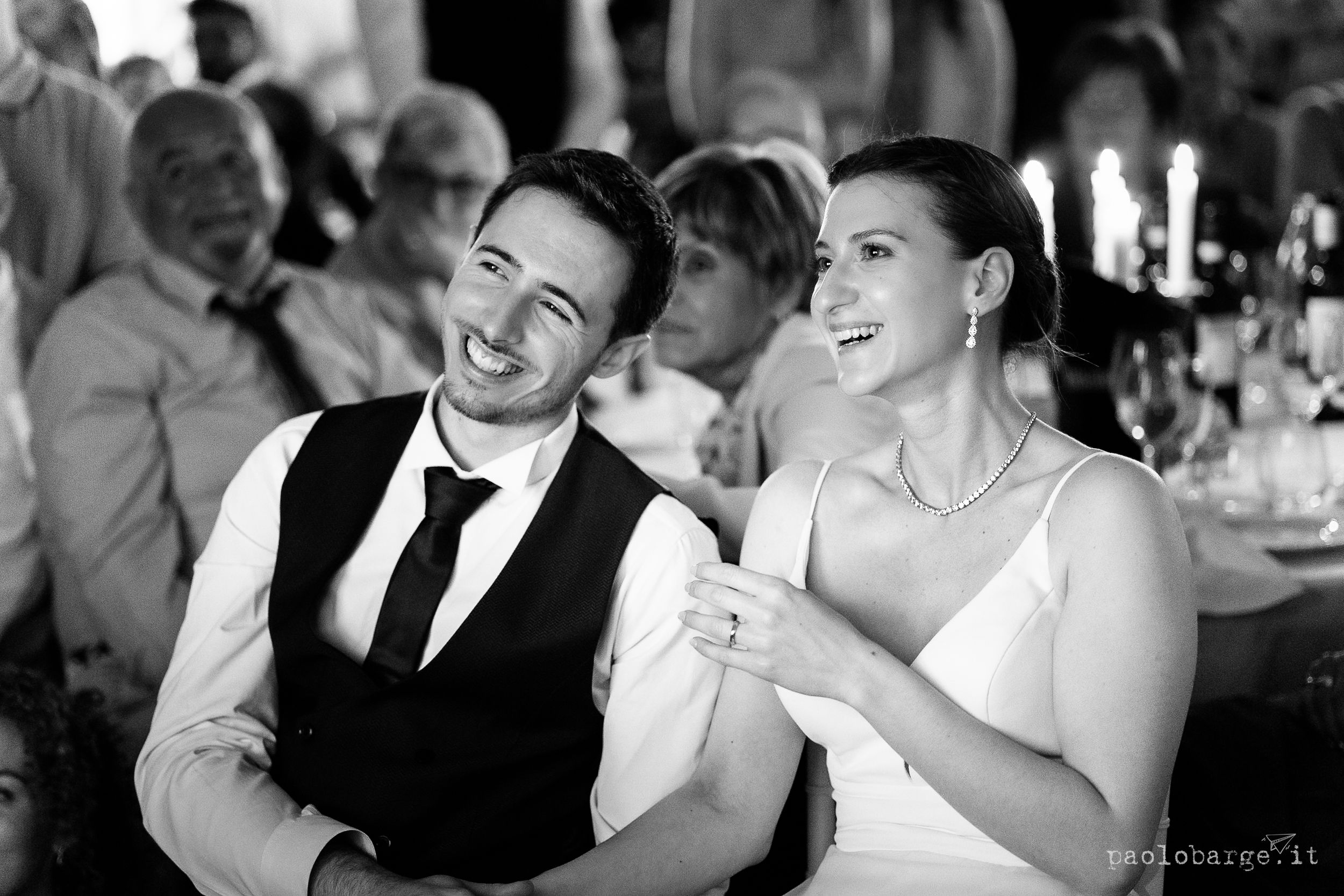 A couple shares a heartfelt laugh together at an indoor reception with candlelit tables in the background.