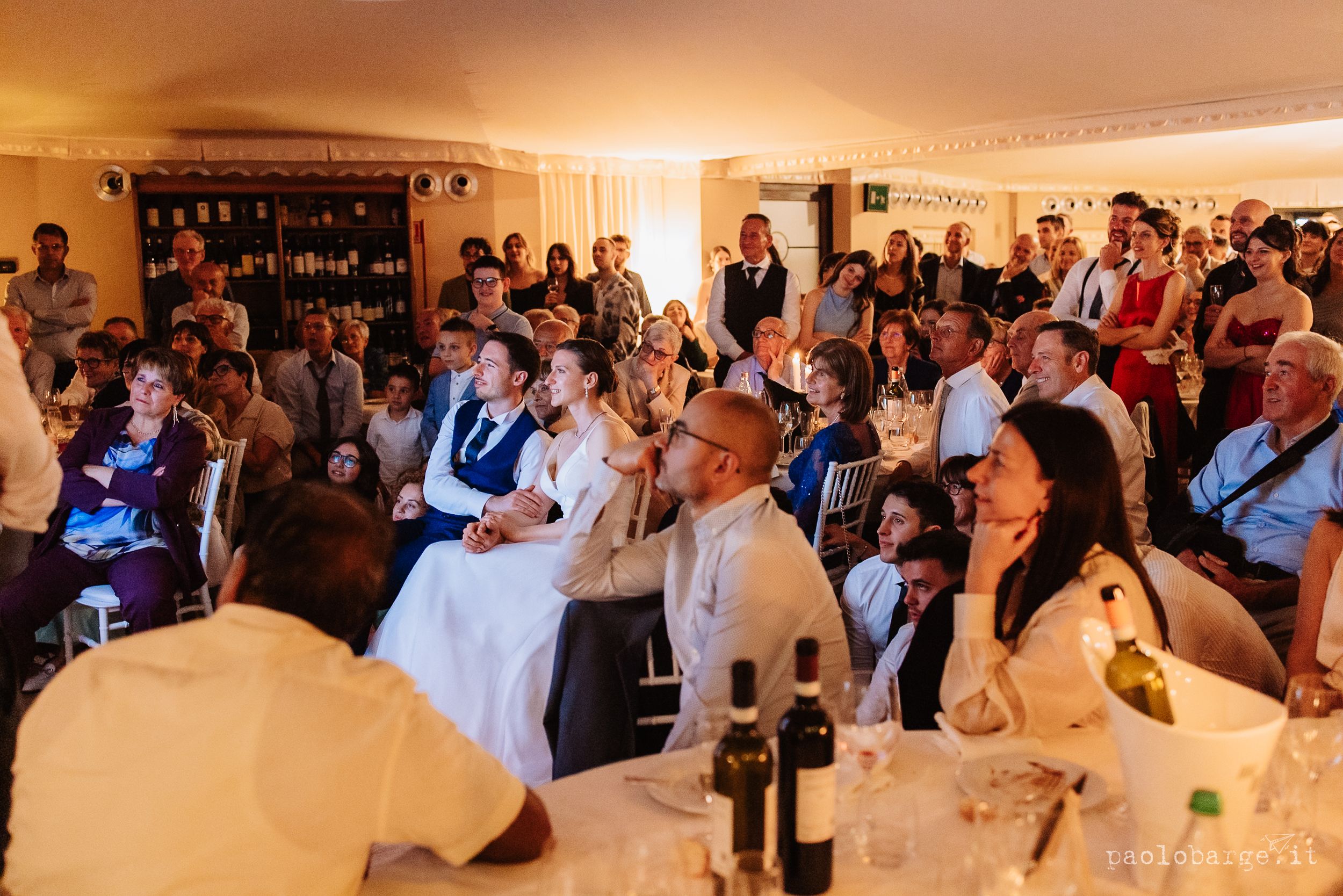 Wedding guests gather at a warm-lit reception venue to watch speeches, with tables set for dinner and wine bottles visible.