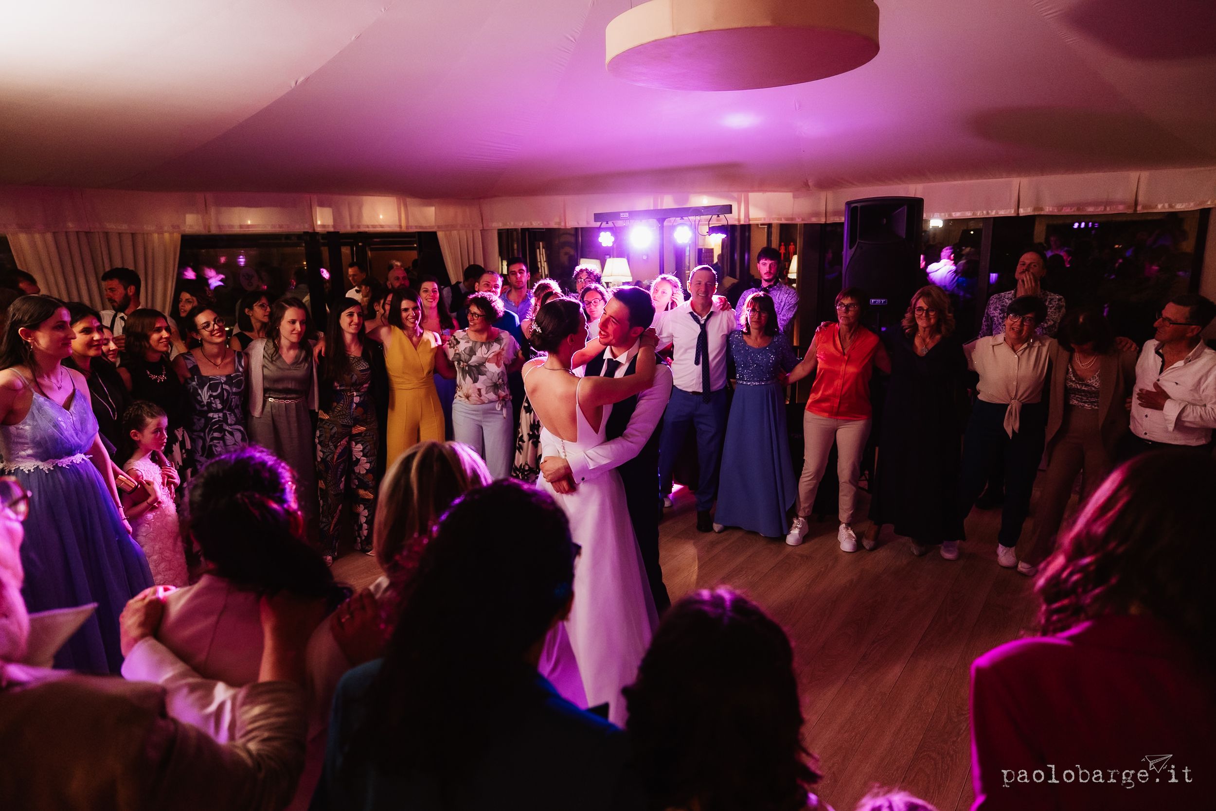 A couple shares their first dance at a wedding reception while guests watch under purple mood lighting in a ballroom.