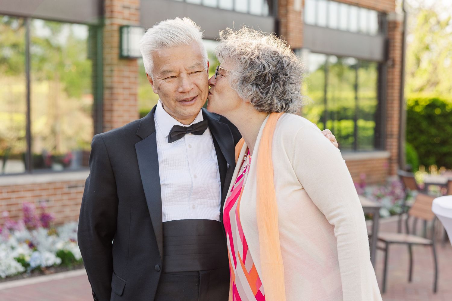 An elegant couple share a tender moment outside on a patio with colorful flowers in the background.