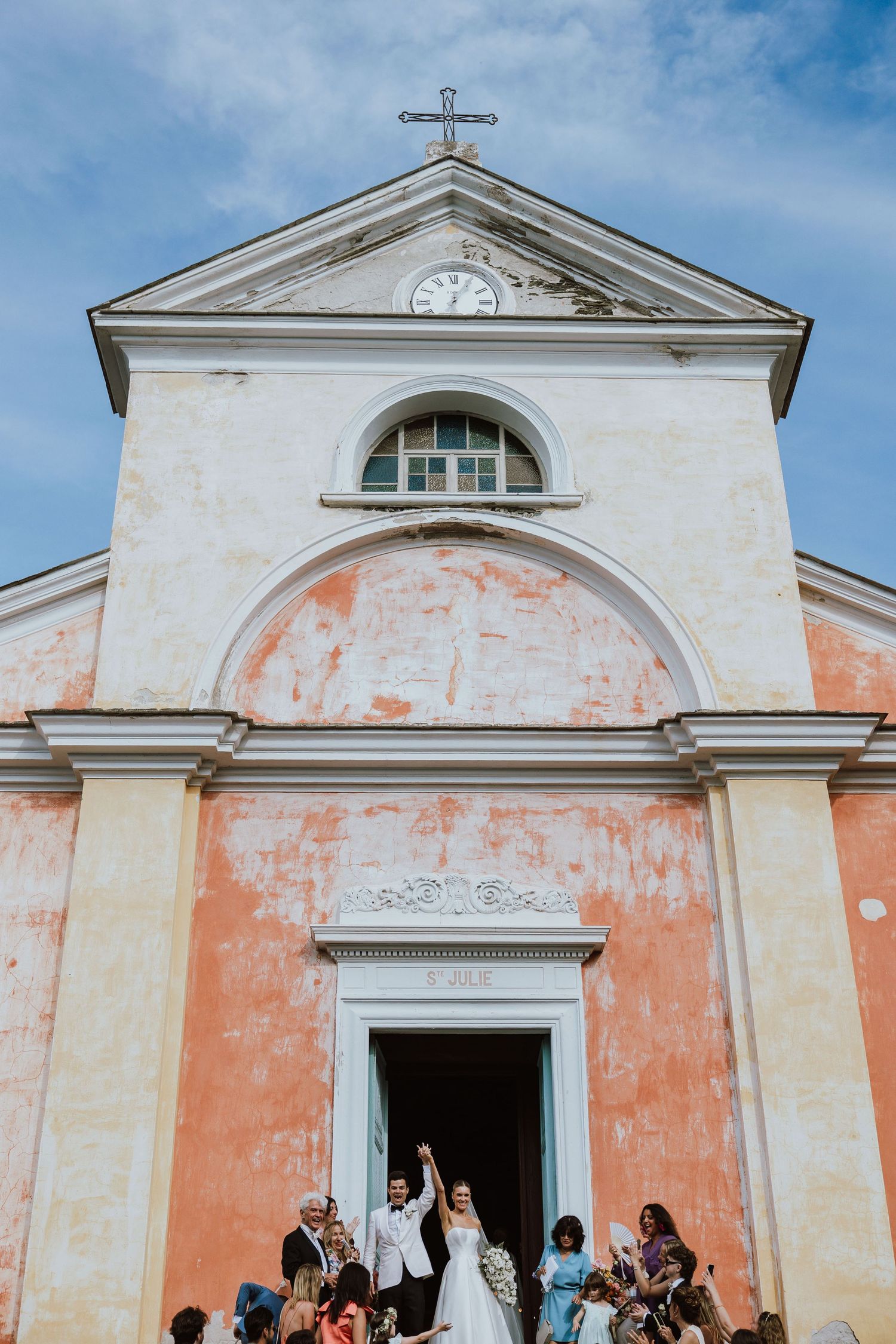 Wedding celebration on church steps with pink and white facade and cross on top against blue sky.