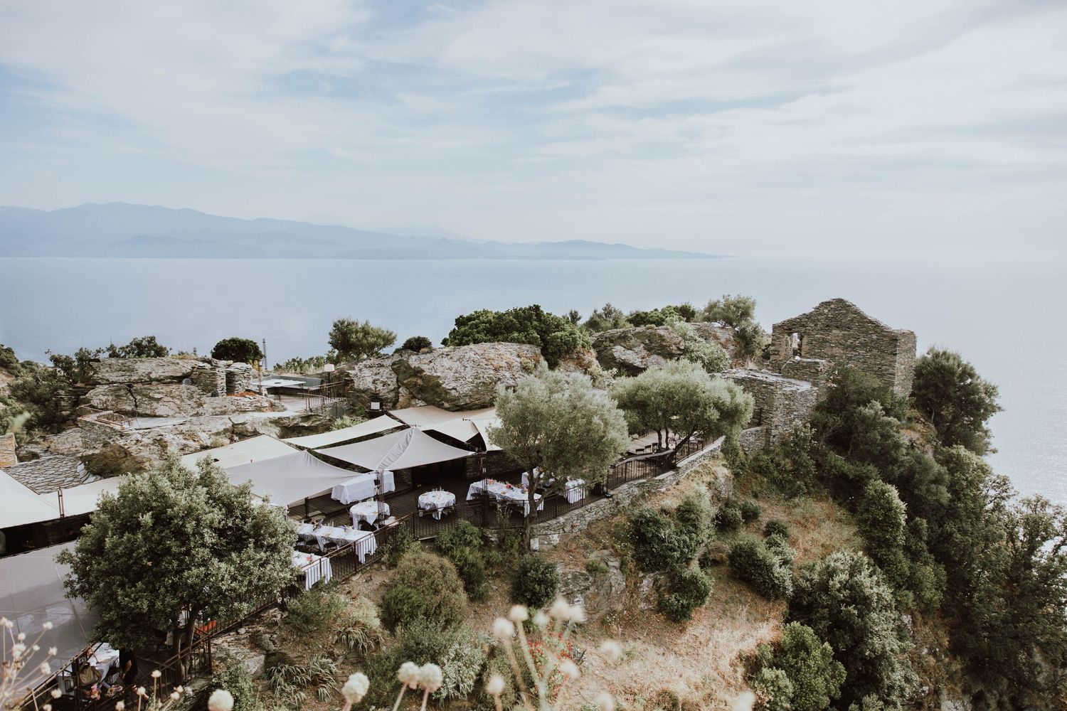 Aerial view of seaside hilltop buildings nestled among trees with calm ocean and mountains in the background.