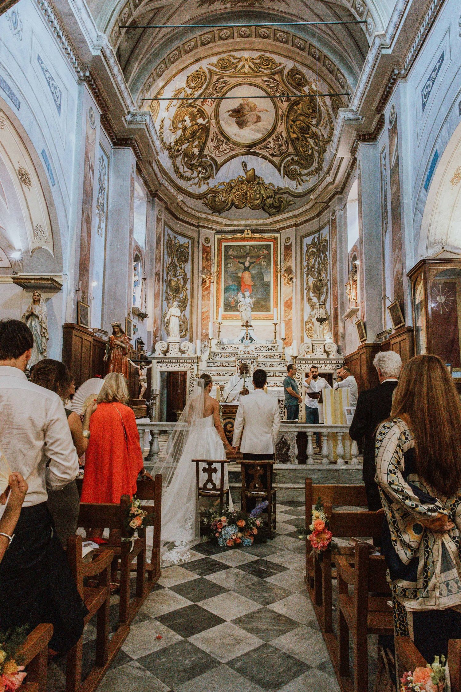 Interior panorama of an ornate Italian church with guests seated in wooden pews and elaborate baroque decorations.
