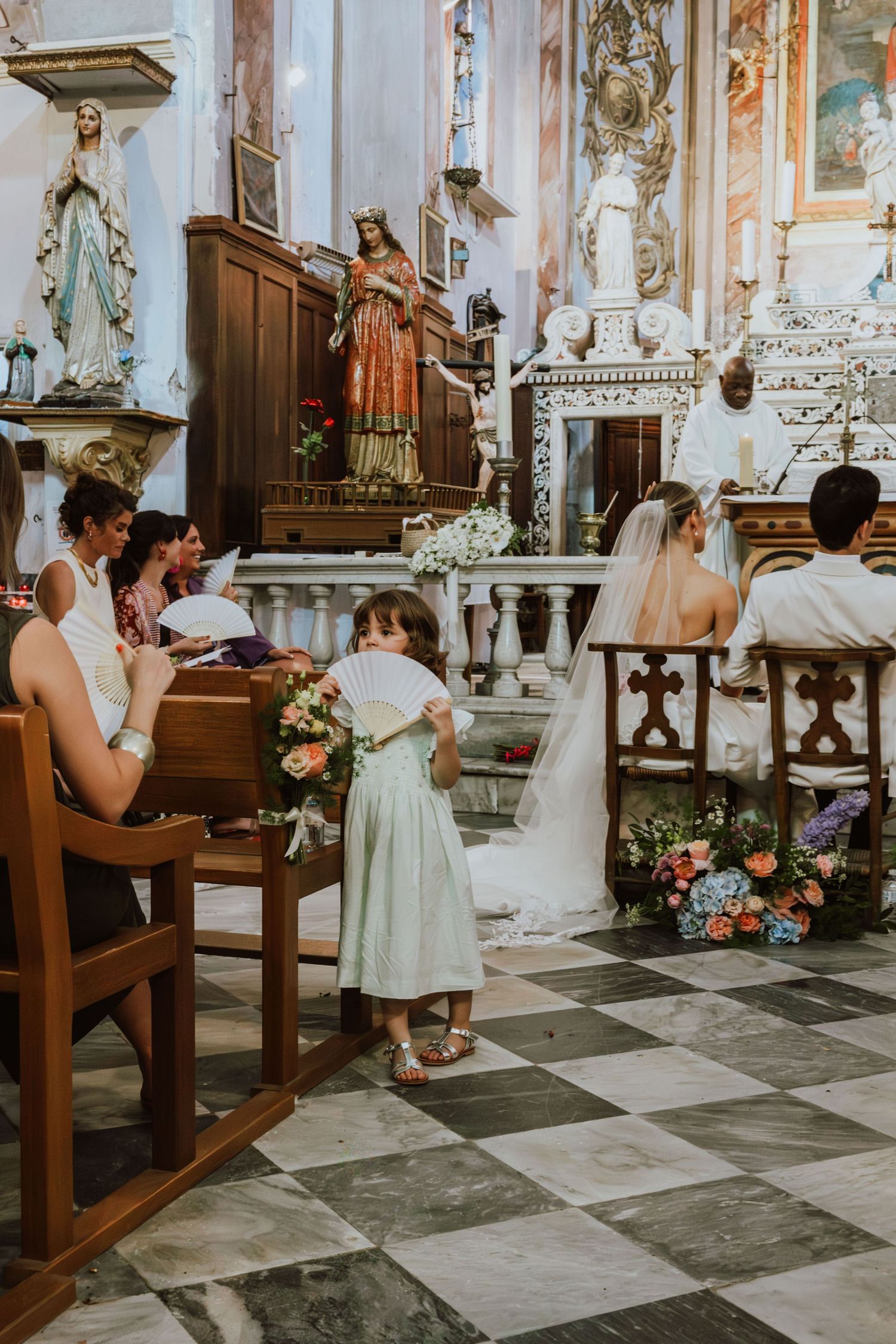 People gather in an ornate church interior with checkerboard floors and statues for a wedding ceremony.