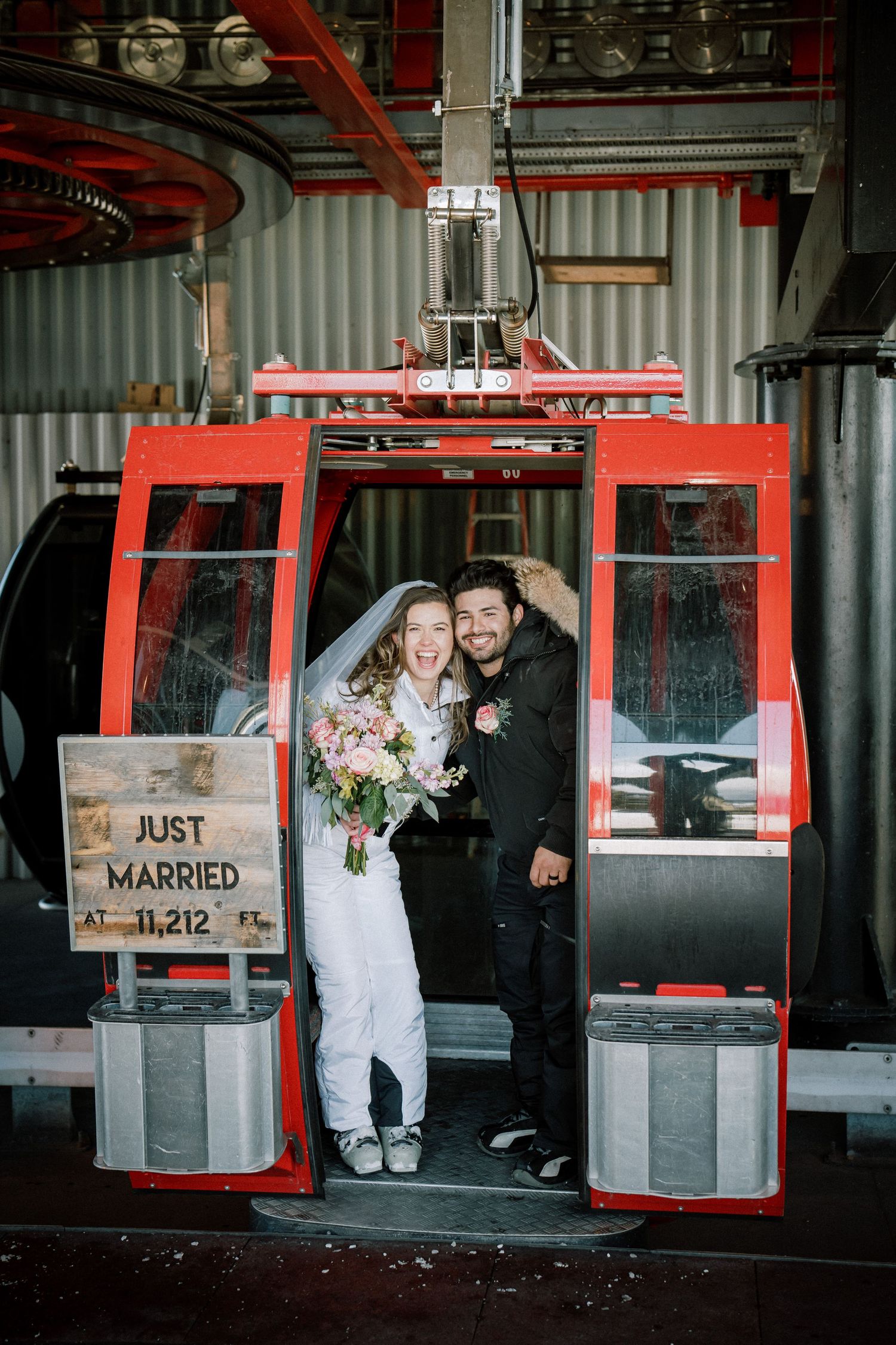 A newlywed couple poses inside a red ski lift gondola with a 'Just Married' sign at their winter wedding venue.