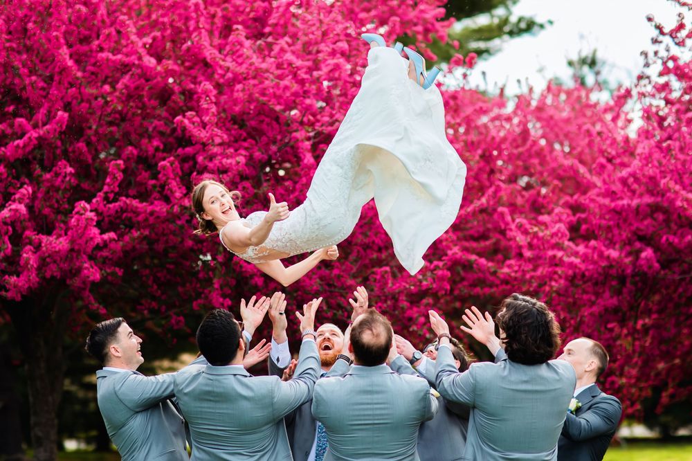 A celebratory scene with light gray suits and flowing white fabric against vibrant magenta flowering bushes.