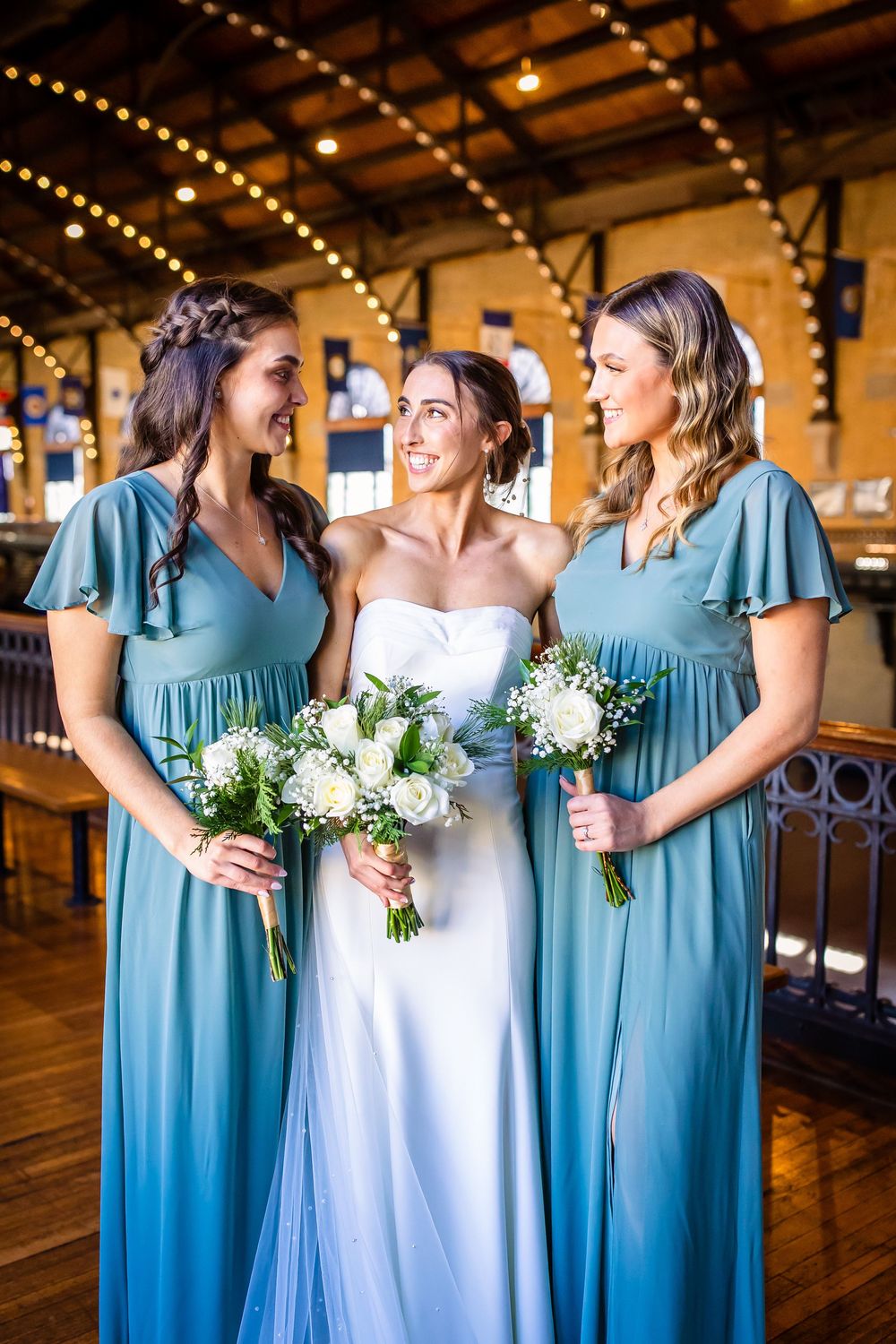 Three women in elegant wedding attire share a joyful moment, with bridesmaids in blue gowns and white floral bouquets.