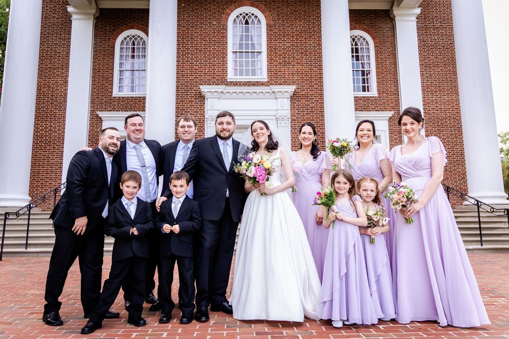 Wedding party poses on brick steps in front of traditional church with bridesmaids in lavender dresses and groomsmen in black.