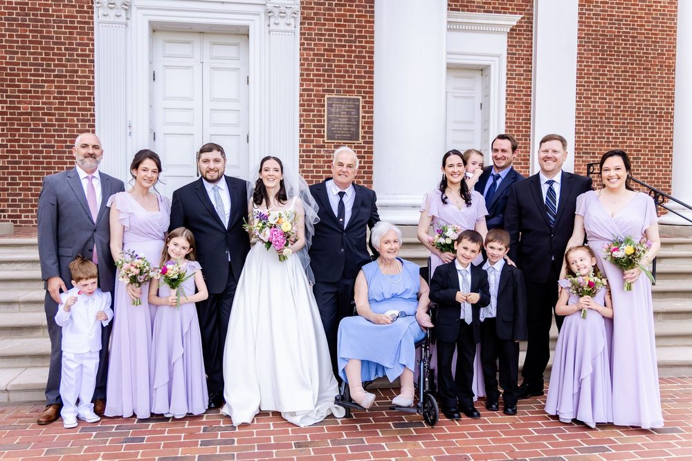 Wedding party stands outside a brick building in lavender bridesmaid dresses and formal attire for group photos.