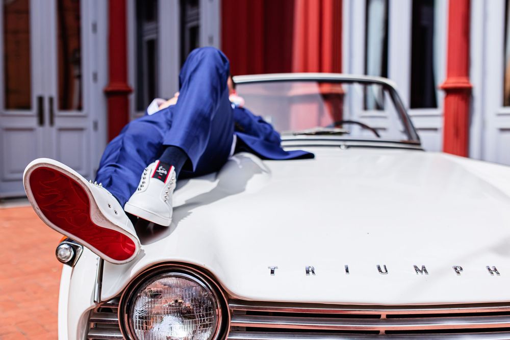 Person in blue hoodie and red sneakers laying across the hood of a vintage white Triumph car.