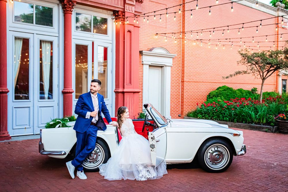 A couple celebrates near a vintage white car parked against a coral colored building with string lights.