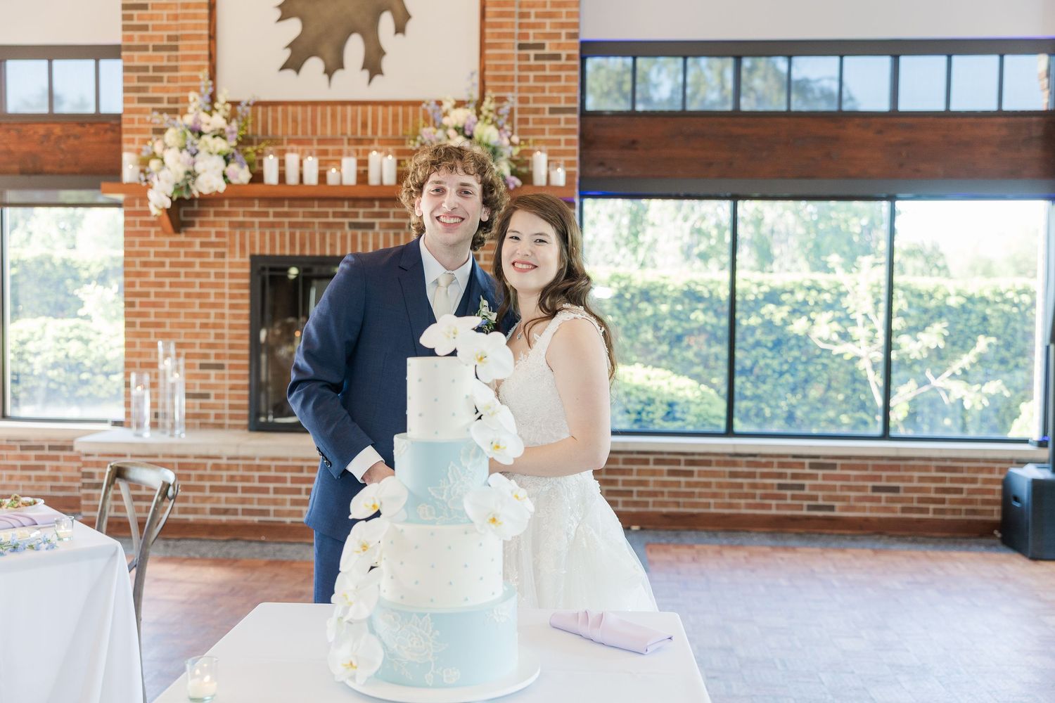 A couple poses with their elegant light blue and white four-tier wedding cake decorated with floral accents at a brick venue.