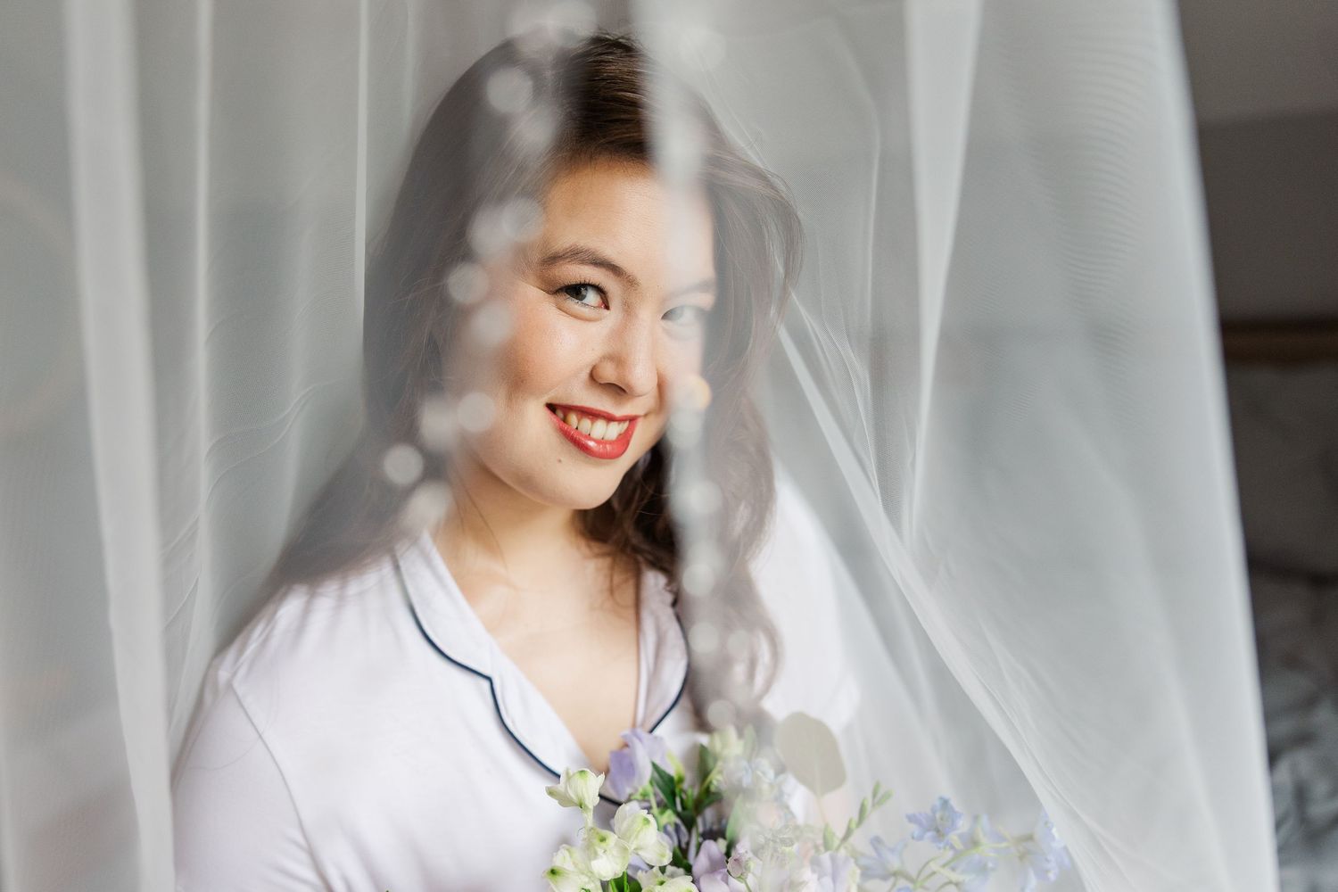 A traditional white wedding gown with a satin bodice and flowing tulle skirt is displayed against sheer curtains.