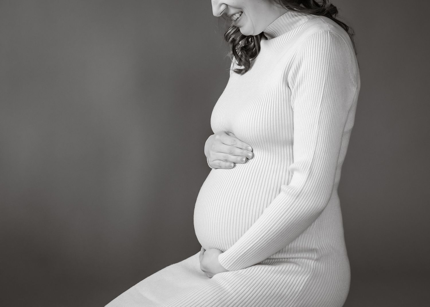 Black and white profile portrait of an expectant mother in a white knit dress holding her baby bump.