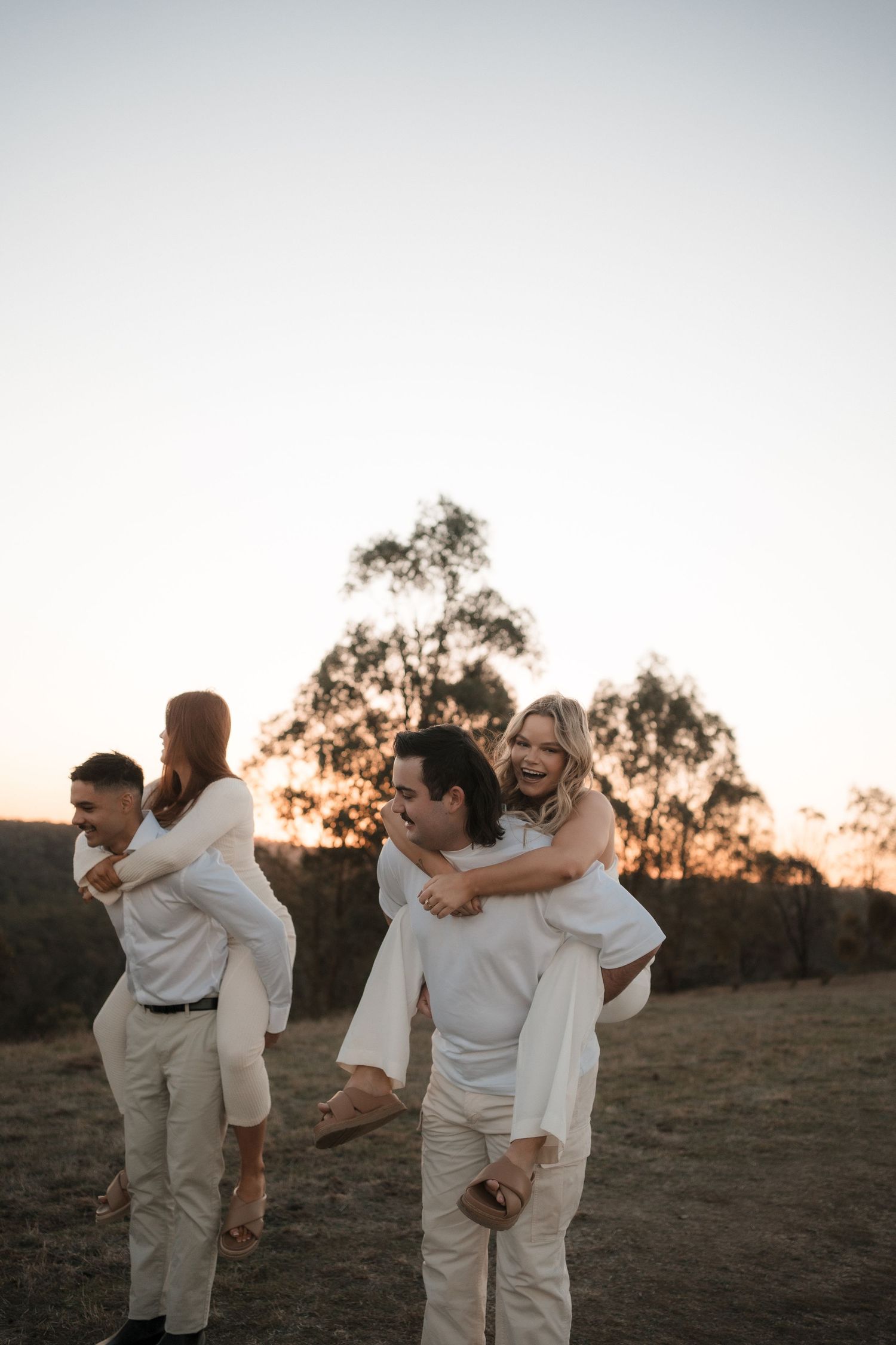 A couple in white outfits playfully embraces and dances together at sunset against a backdrop of silhouetted trees at Mt Lofty, Wonga Park