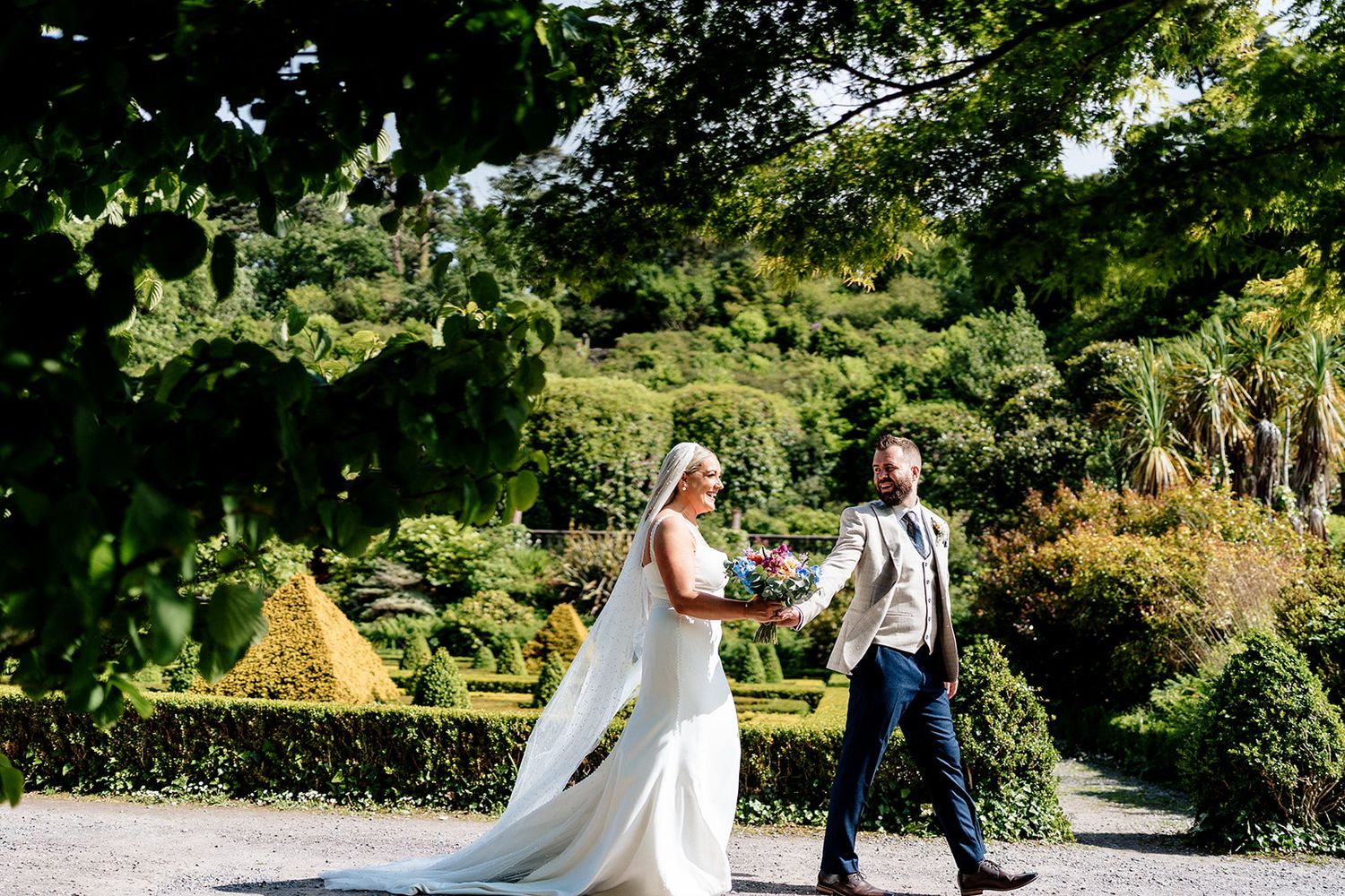A couple celebrates their wedding day walking together along a sunny garden path at an outdoor venue.