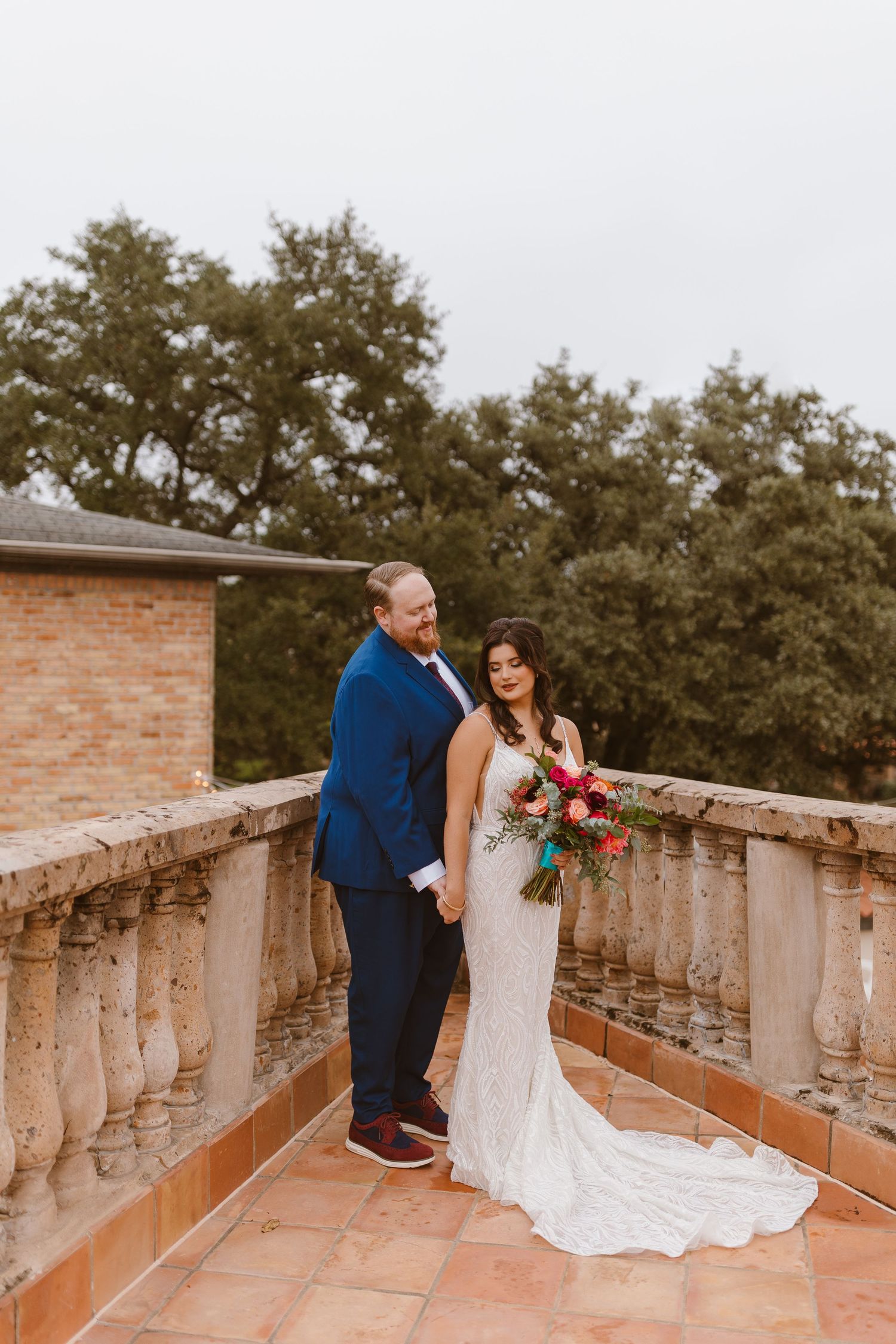 Couple in formal wedding attire standing on a terracotta tile balcony with stone balustrades and trees in background.