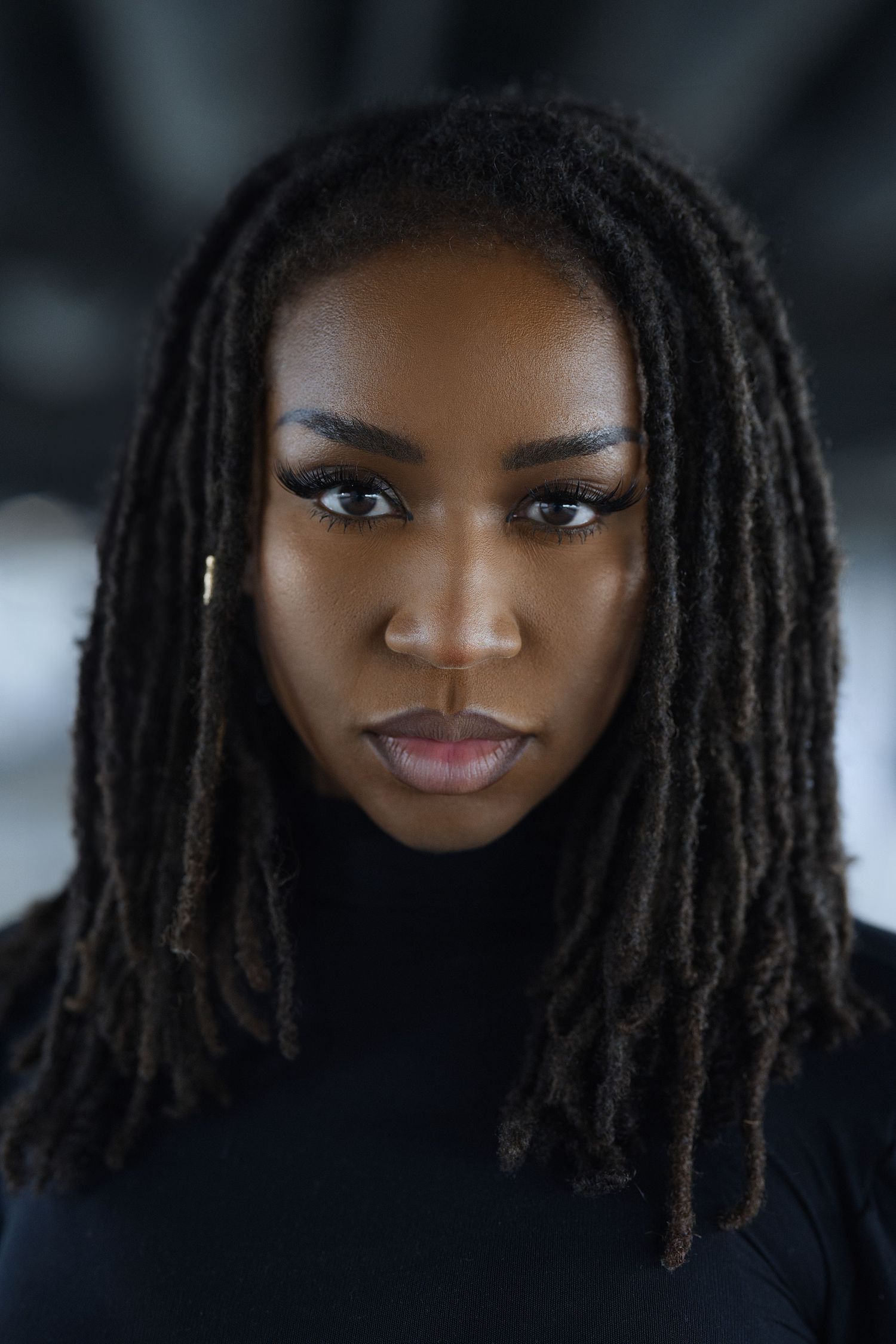 Jordan Frazier: Professional portrait of a person with long locs wearing a black turtleneck against a dark background.