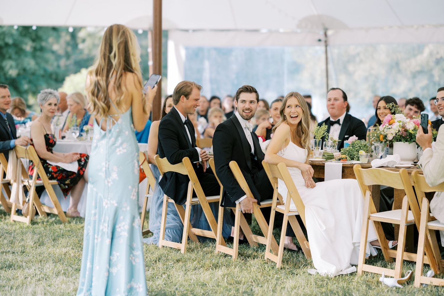 Guests seated at long outdoor wedding reception tables under a tent with blue mountains visible in background.