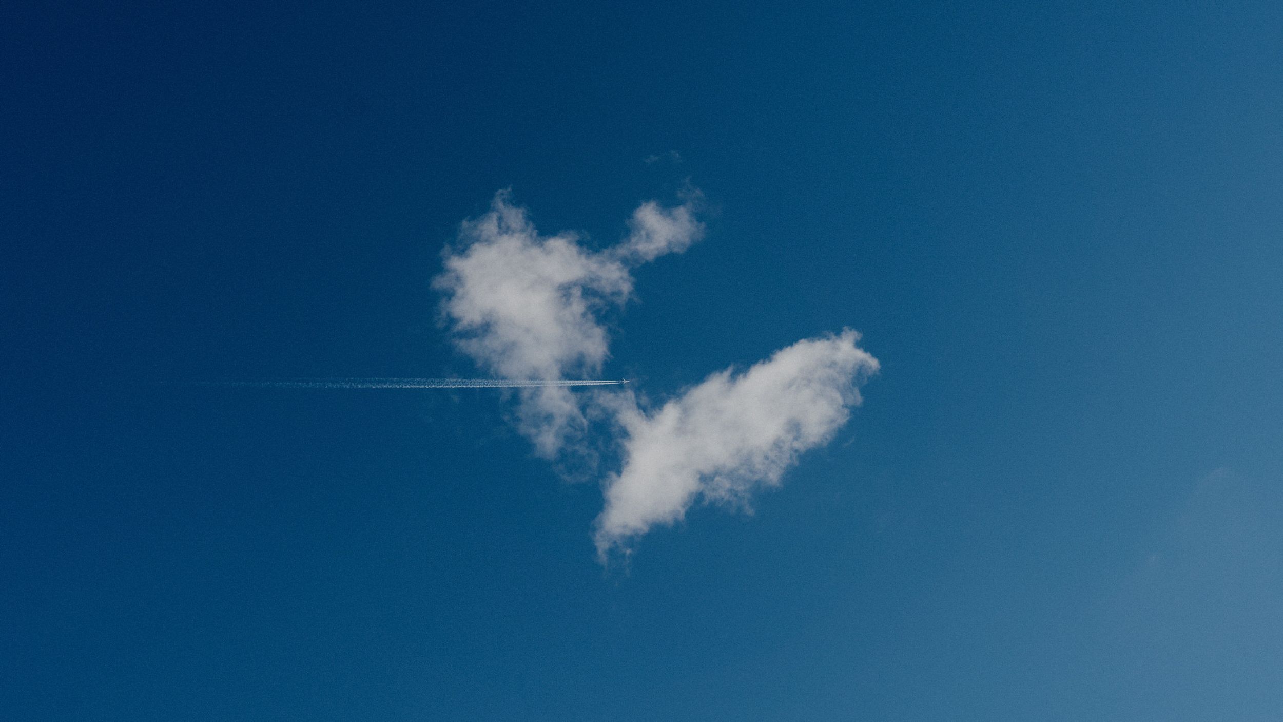 A lone white cloud formation against vibrant blue sky with a thin airplane contrail streaking across.