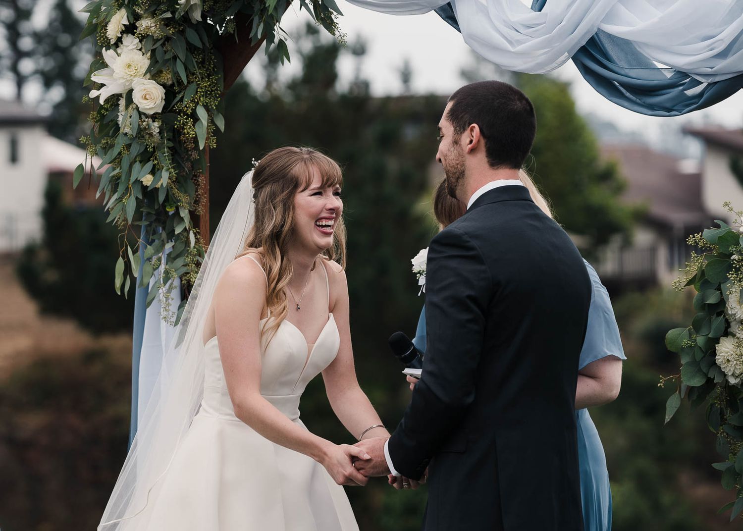 Wedding couple shares a joyful moment under a floral arch decorated with white flowers and greenery.