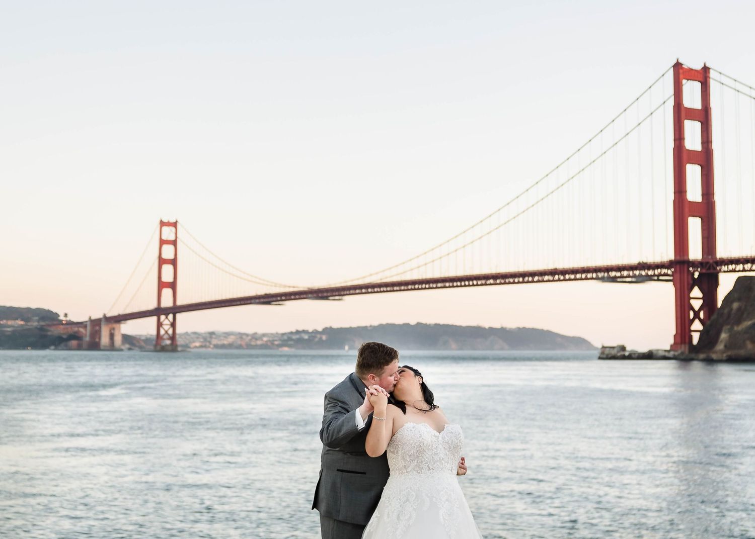 Elegant wedding scene with couple embracing by the iconic Golden Gate Bridge in San Francisco at sunset.
