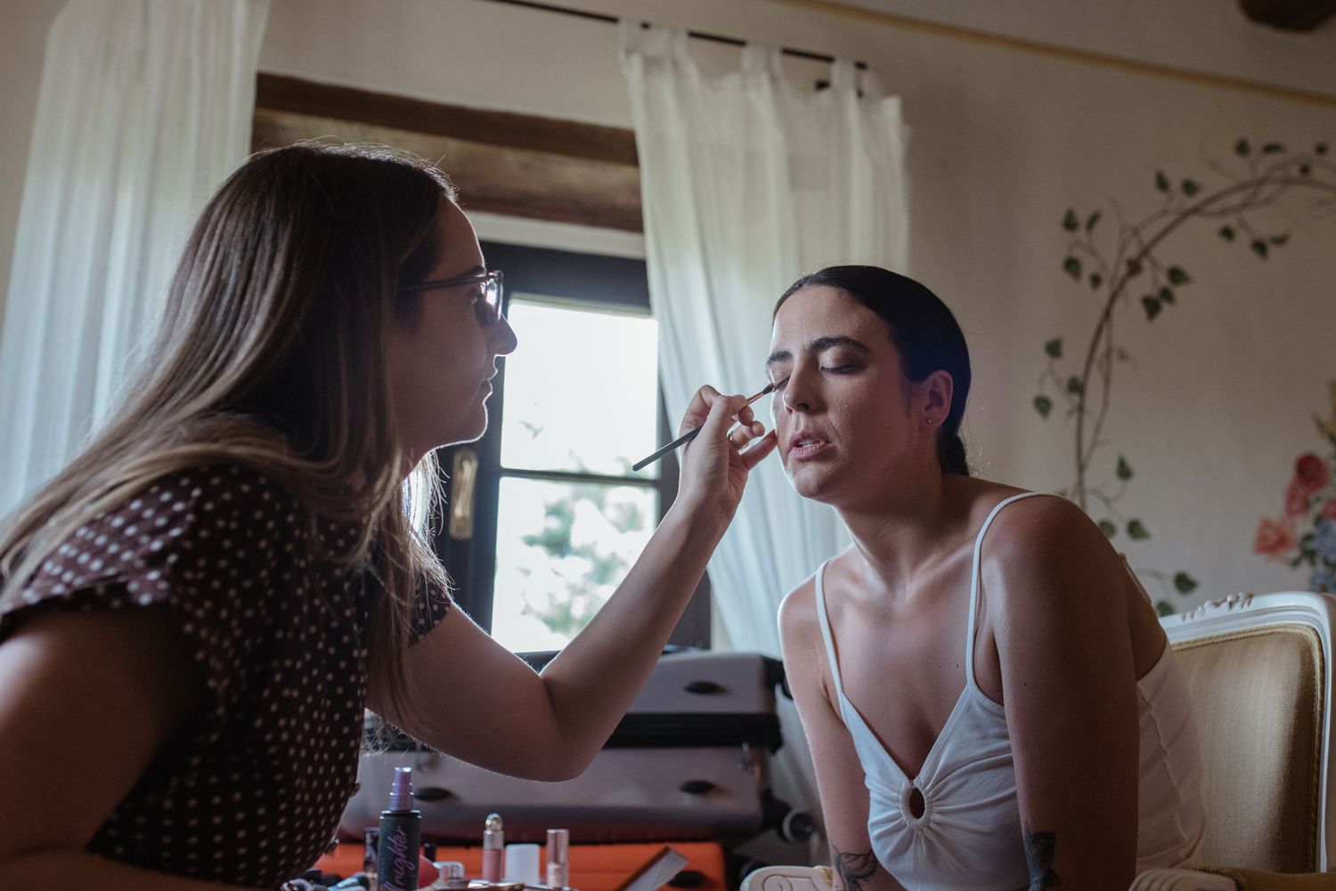 Makeup artist applies cosmetics to client sitting in soft natural window light in home setting.