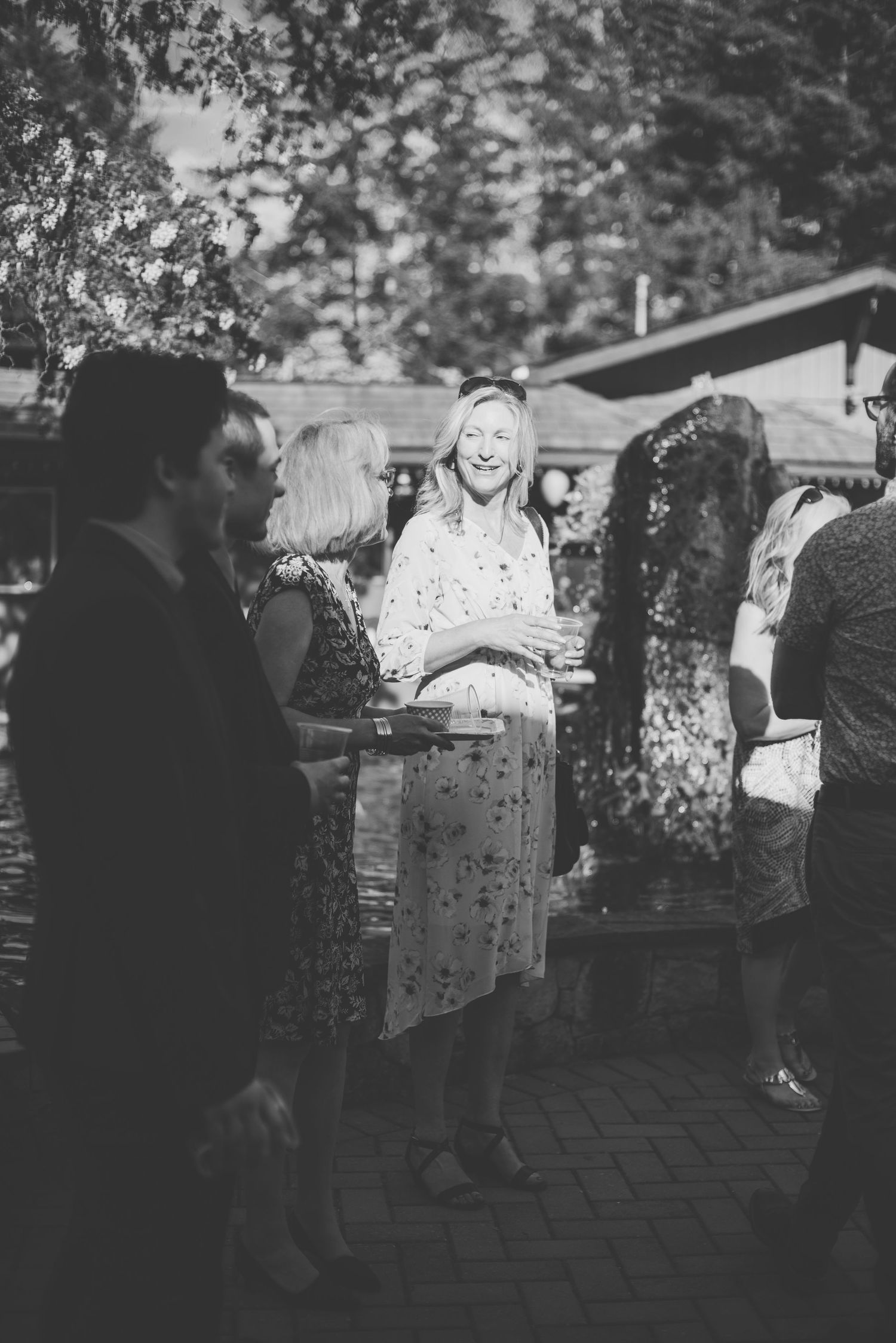 A black and white candid shot of people gathered at an outdoor social event with dappled lighting through trees.
