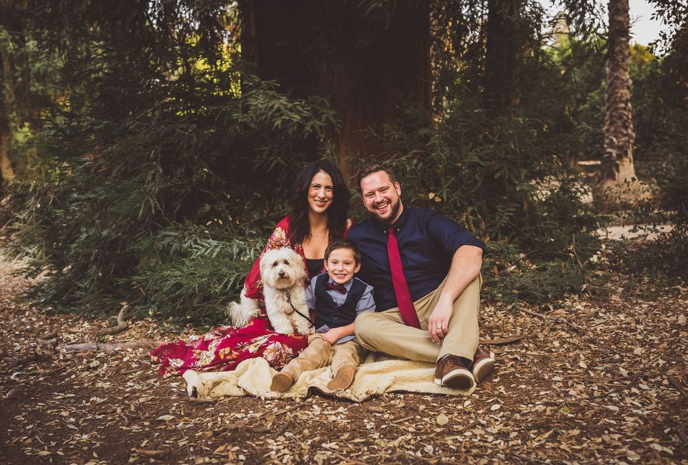 A family sits together on a red blanket with their fluffy white dog during an outdoor photo session in a wooded setting.