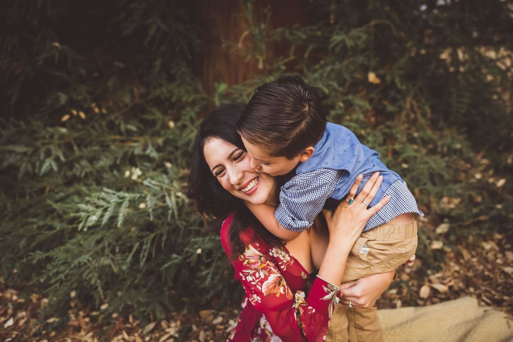 A couple shares intimate moments in a forest setting while wearing burgundy and beige outfits during an engagement photoshoot.