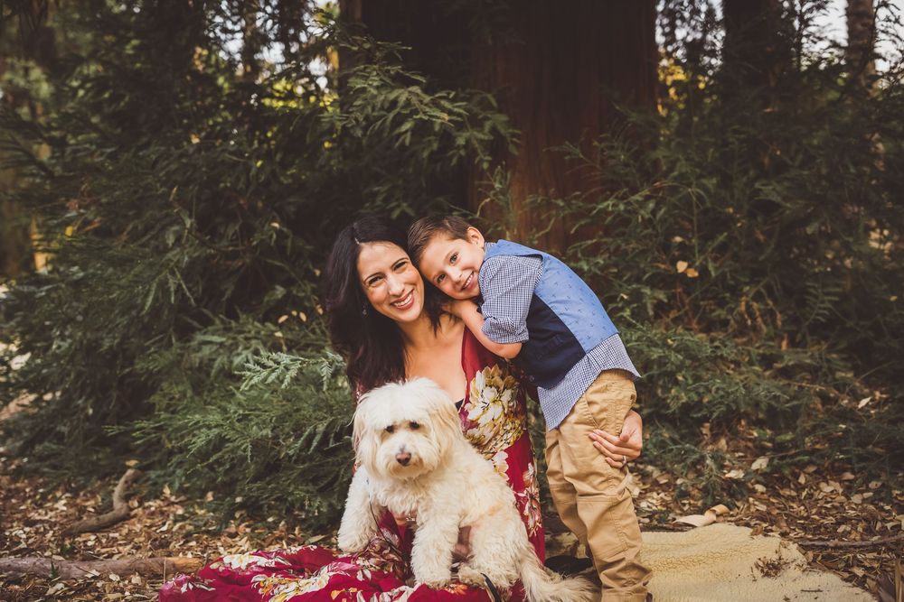 A joyful couple sitting on a red blanket with their fluffy white dog in a scenic autumn forest setting.