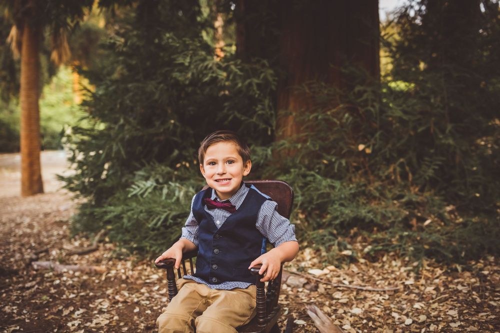 A young student in a navy vest sits outdoors on a forest trail surrounded by trees during autumn.