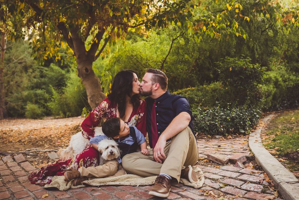 Couple in burgundy colored clothing sits on garden path with small white dog during autumn afternoon.