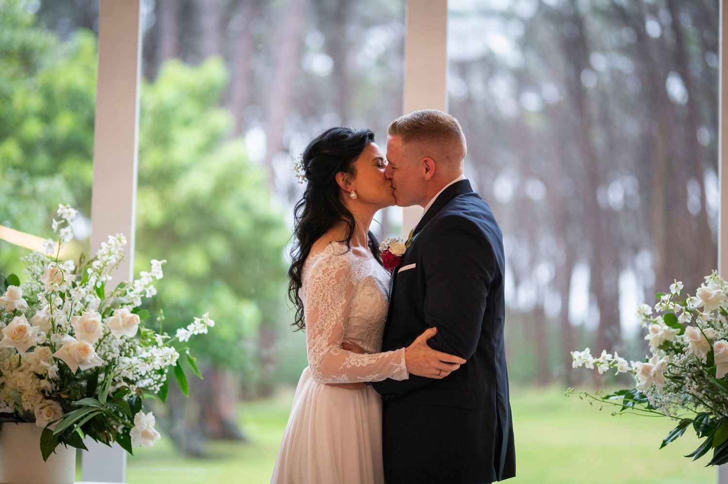 An intimate wedding couple shares a romantic moment in a lush outdoor garden setting with white floral arrangements.