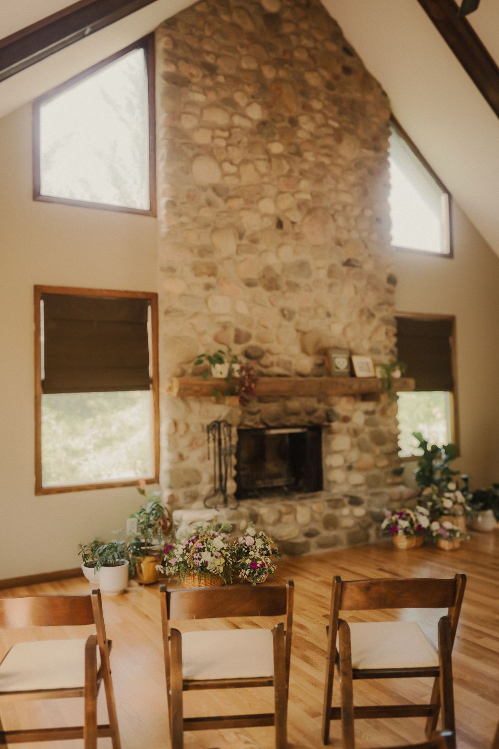 A rustic interior space with stone fireplace, wooden chairs, and natural light streaming through angled windows.