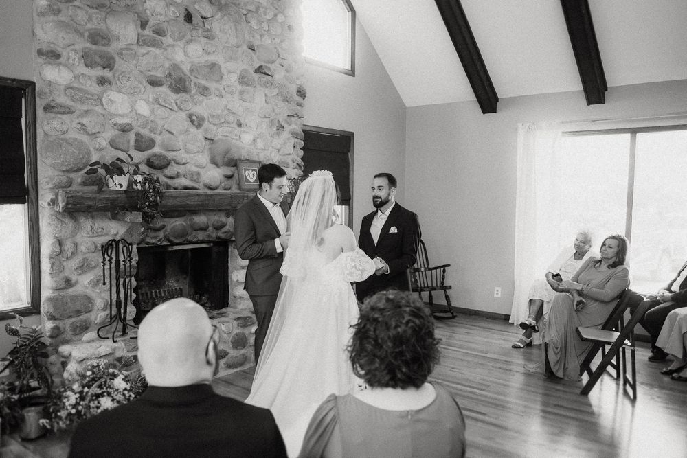 A wedding ceremony takes place in a rustic stone-walled room with exposed wooden beams and guests seated.