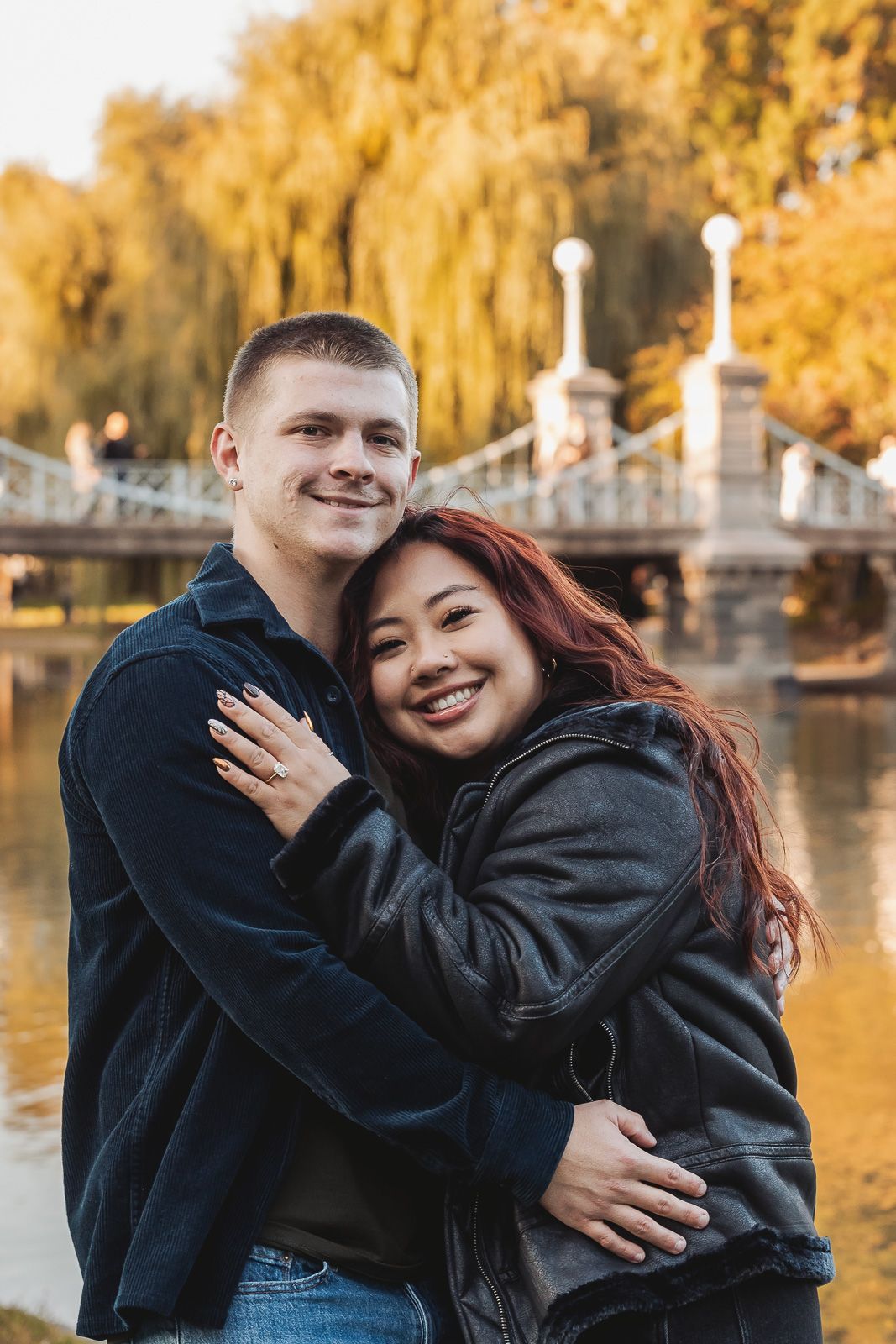 Engaged couple showing ring during autumn photoshoot by ornate bridge and willow trees.