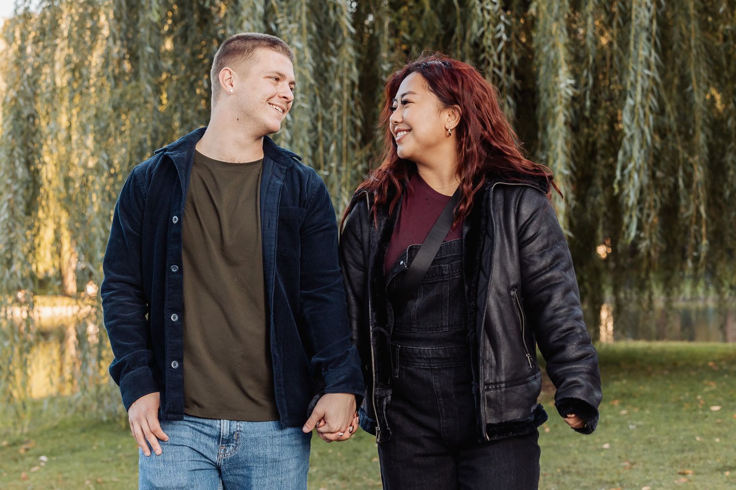 Couple holding hands and laughing under weeping willow trees in a park setting.