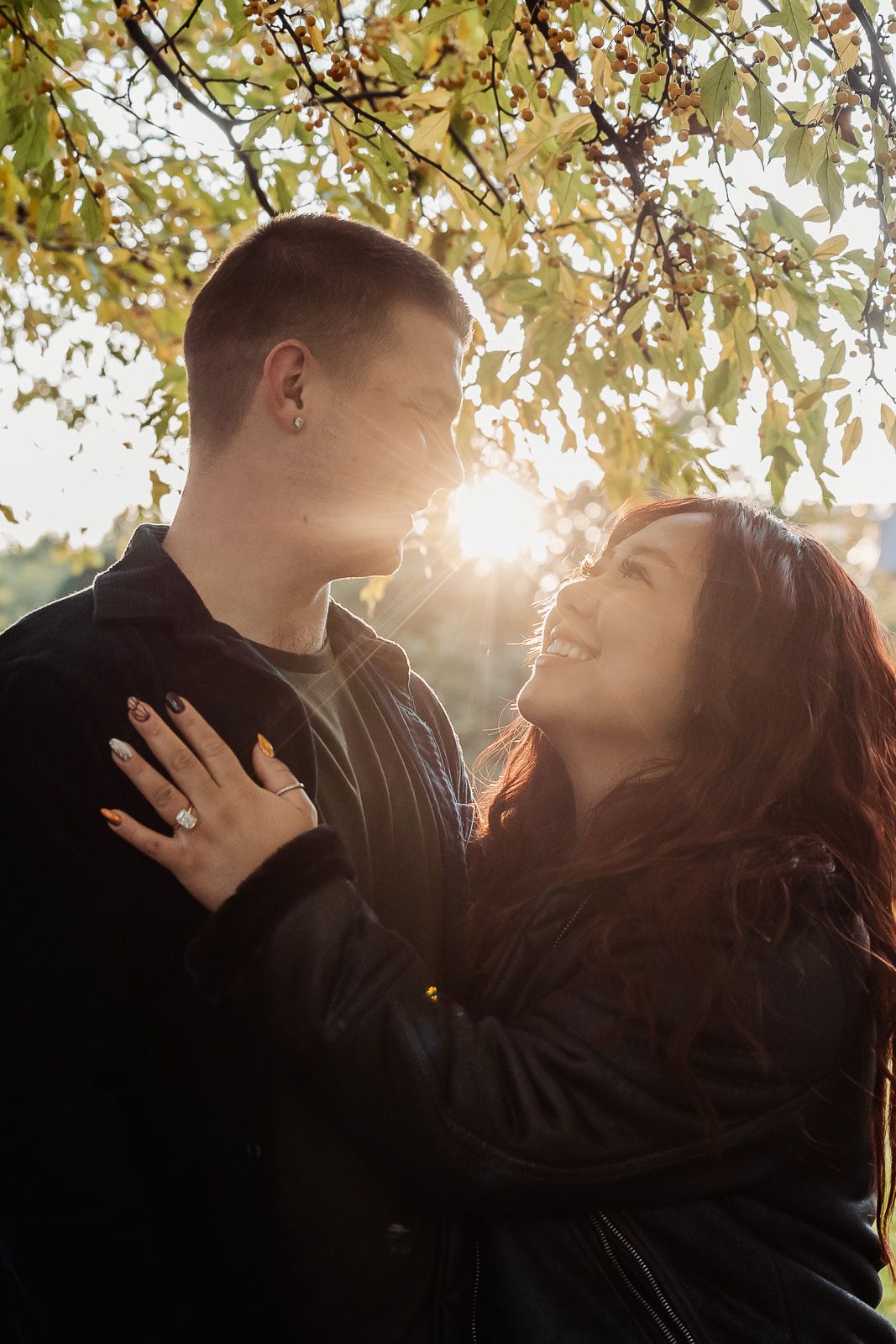 Romantic couple pose in sunlit park with autumn leaves and golden backlight.