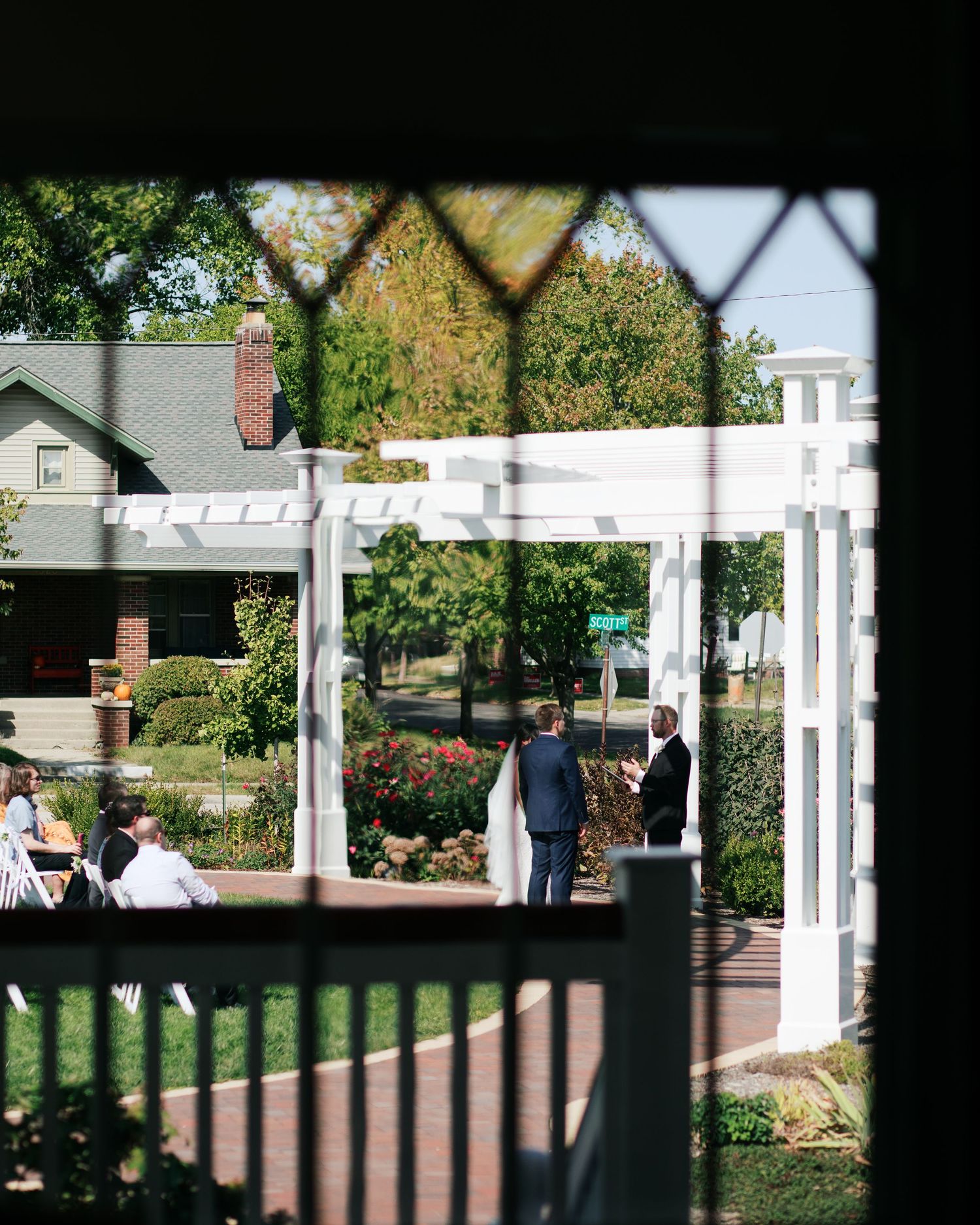 Garden wedding venue with white pergola and rose bushes viewed through decorative window frame.