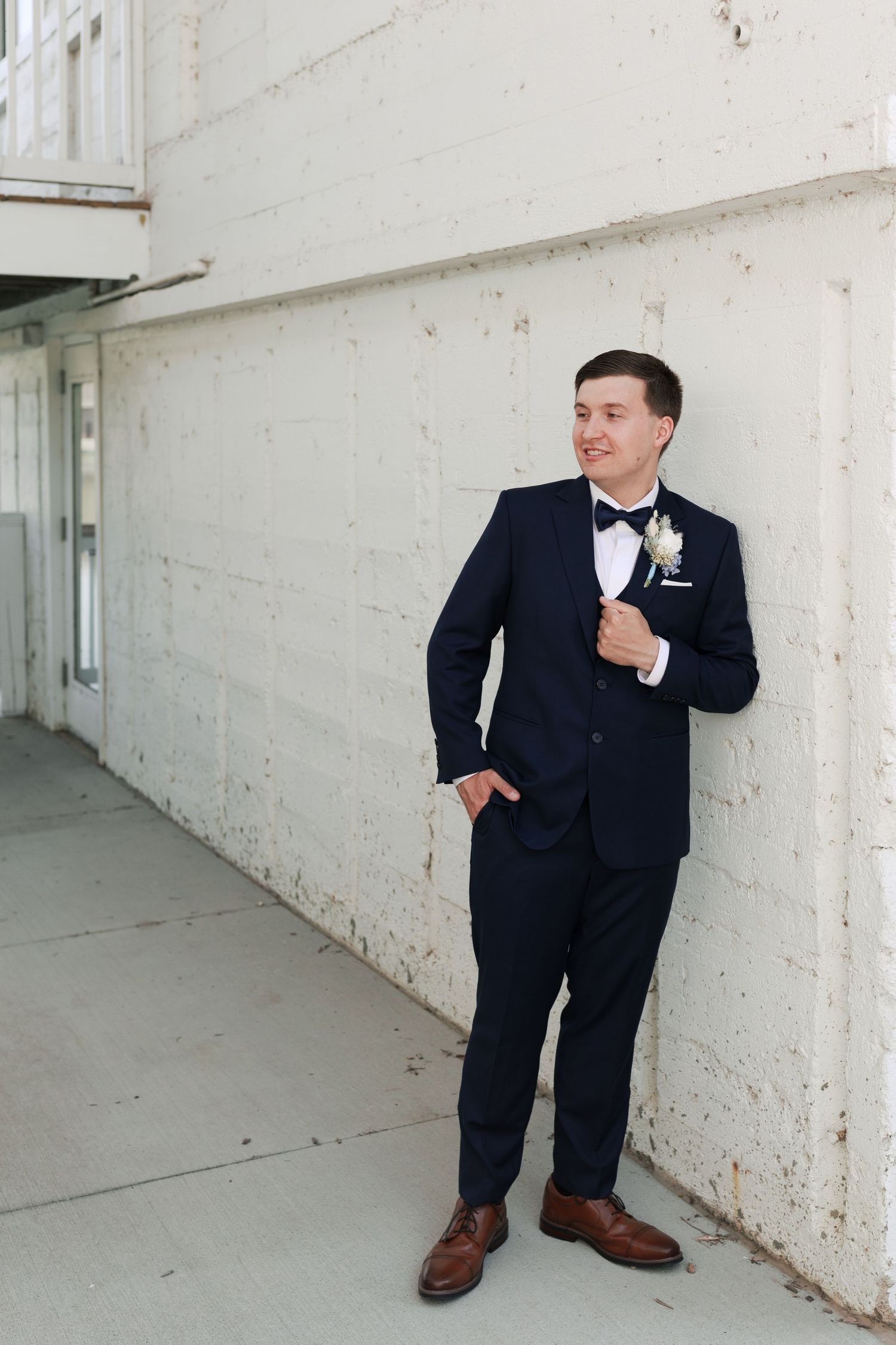 Groom in navy tuxedo poses against white wall with boutonniere.