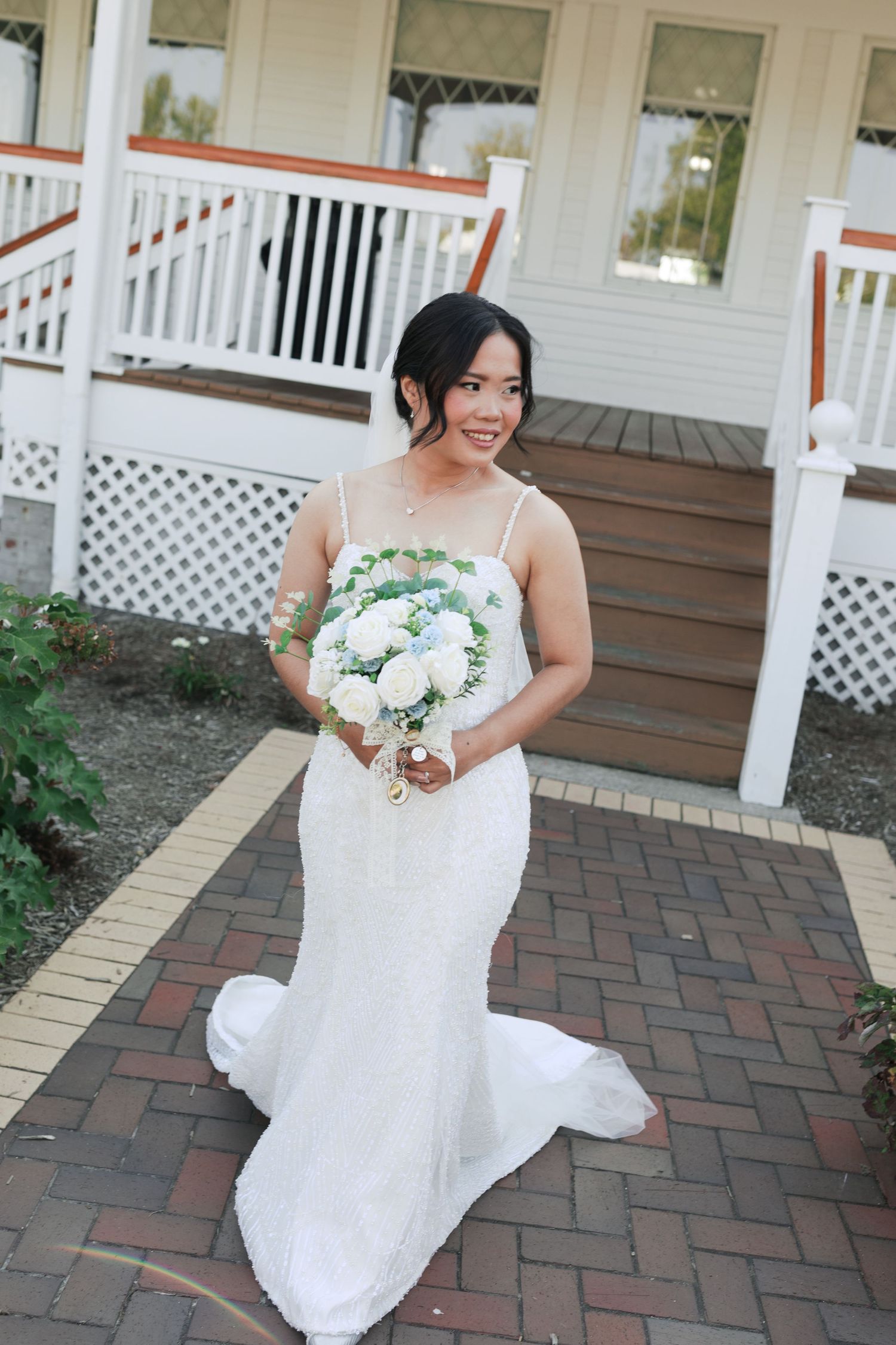 Bride holding white bouquet stands on brick pathway in front of white porch railing.