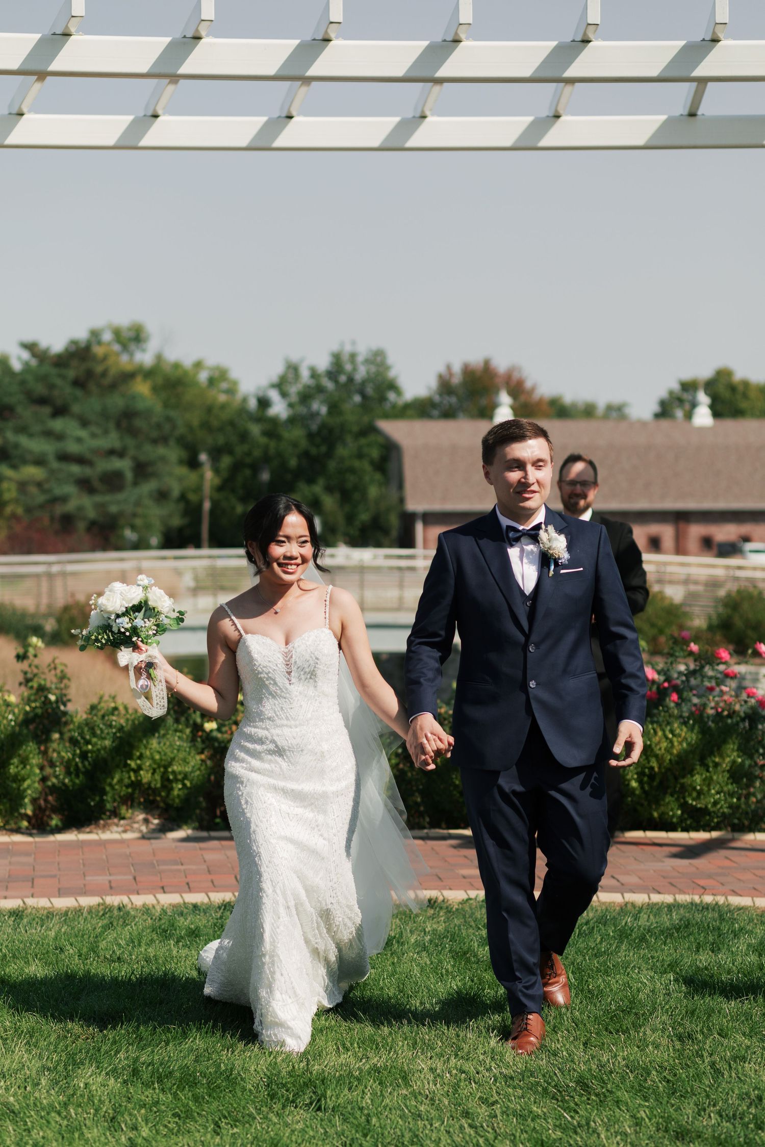 A wedding couple walking together on a brick path under a pergola in a garden setting.