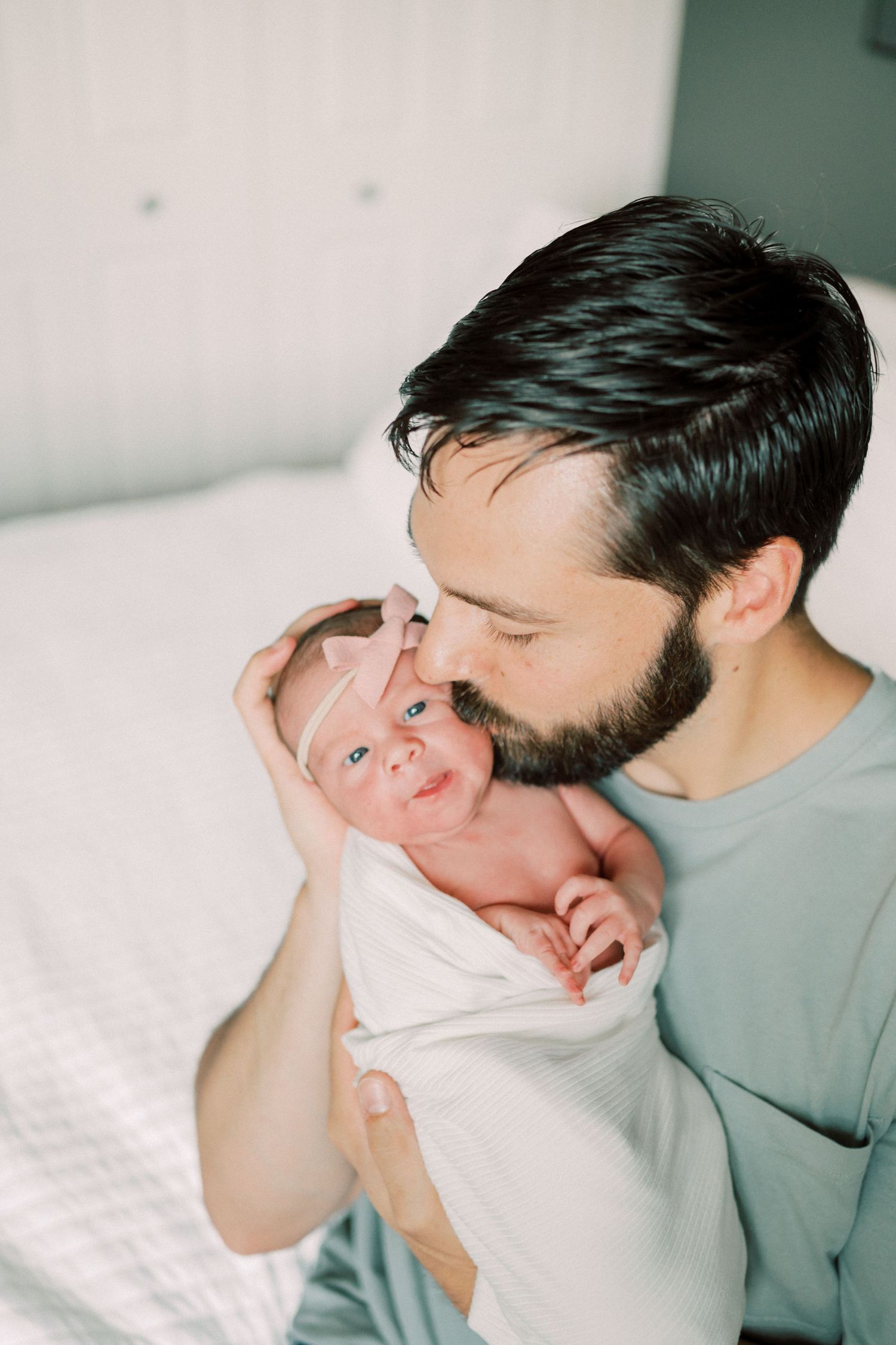 A tender moment between a bearded parent in a grey shirt cradling a tiny newborn baby wrapped in white.