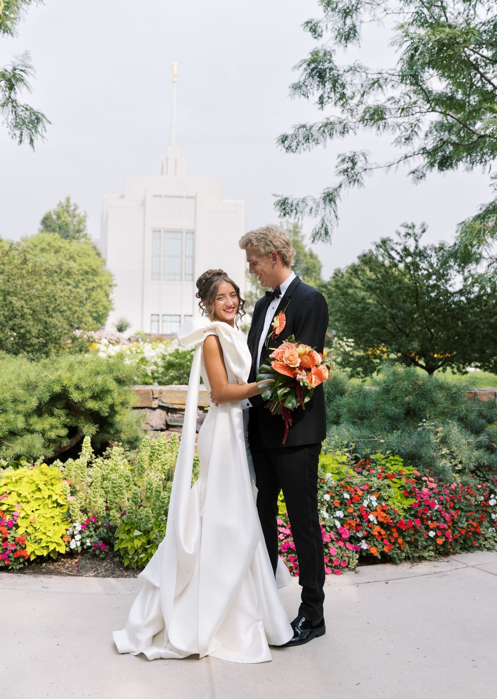 A bride and groom share a romantic moment in a garden with colorful flowers and a church spire visible in the background.
